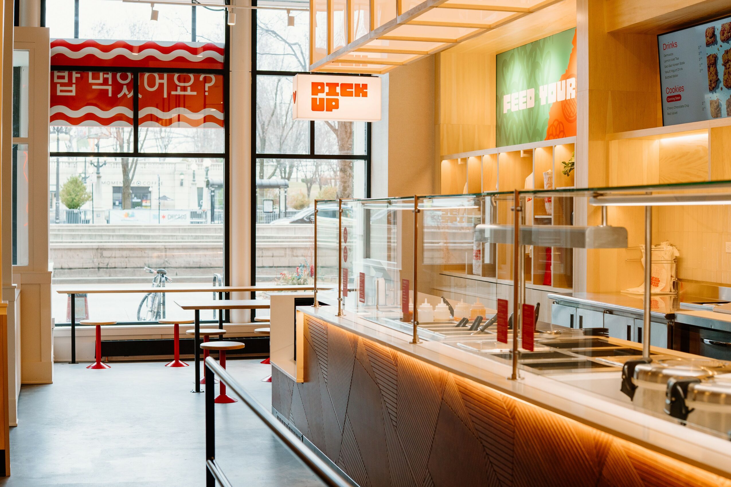 Interior of a modern fast-food restaurant with a counter, glass barrier, and digital menu screens, illuminated by warm lighting.