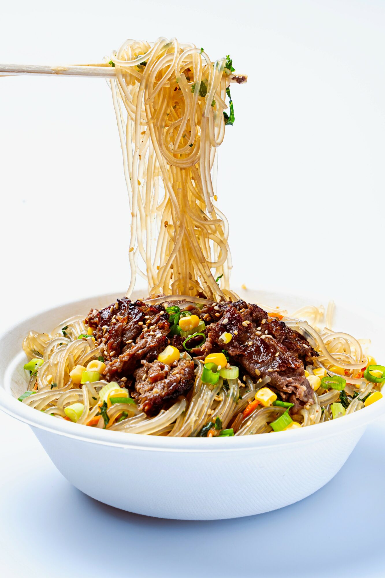 Bowl of noodles with beef and green onions, with chopsticks lifting noodles, on a white background.