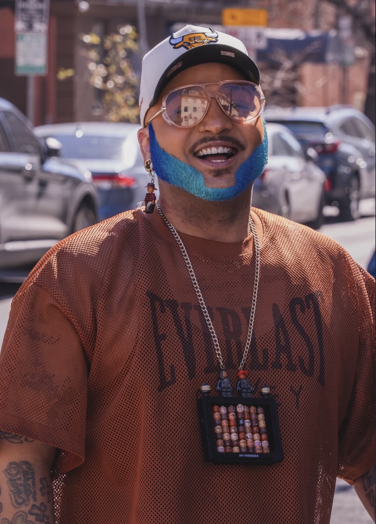 Person with colorful beard, sunglasses, cap, and brown shirt outdoors, smiling, with cars and buildings in background.