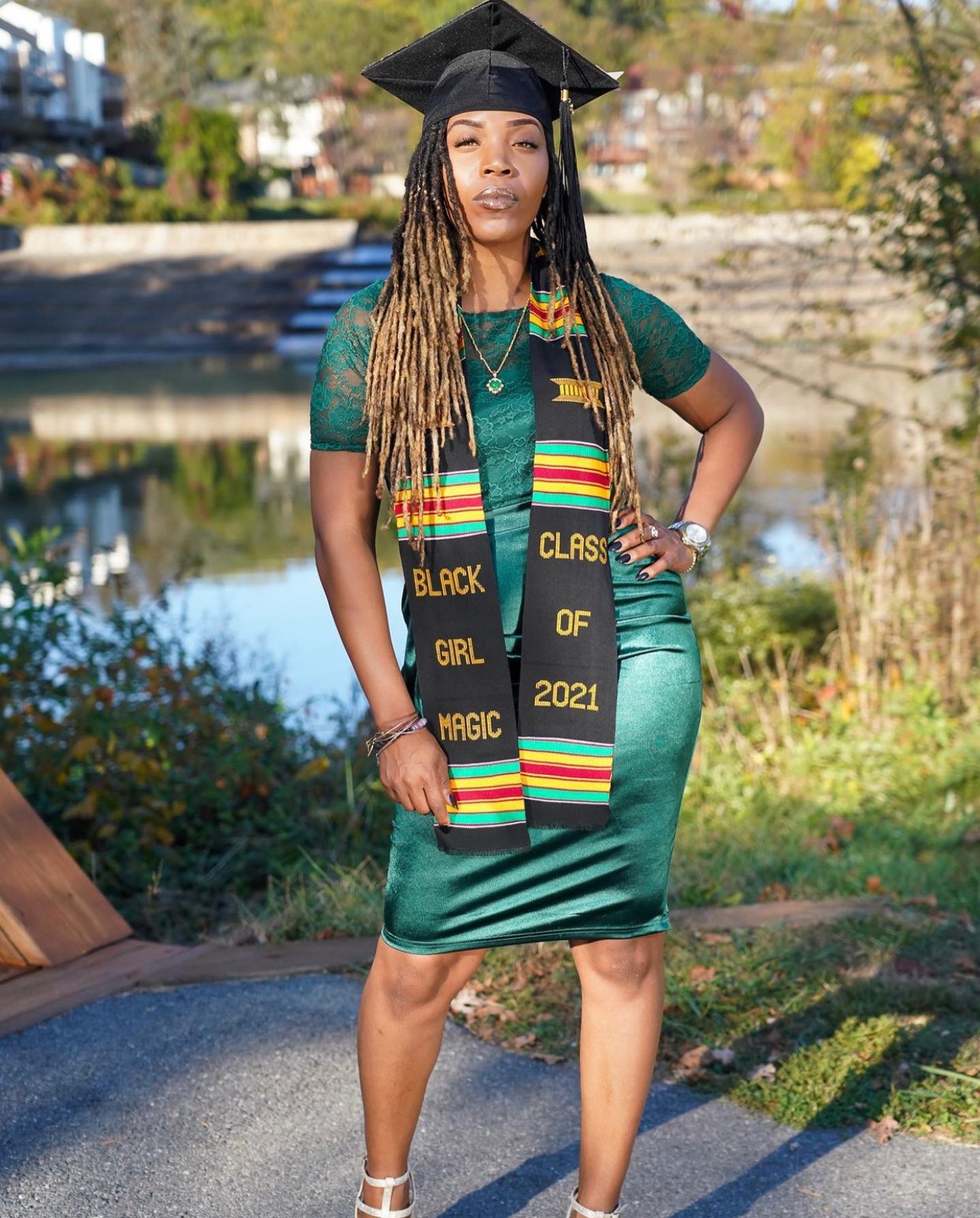 Woman in graduation cap and stole, standing outdoors near water, wearing a teal dress, with trees and a bridge in background.