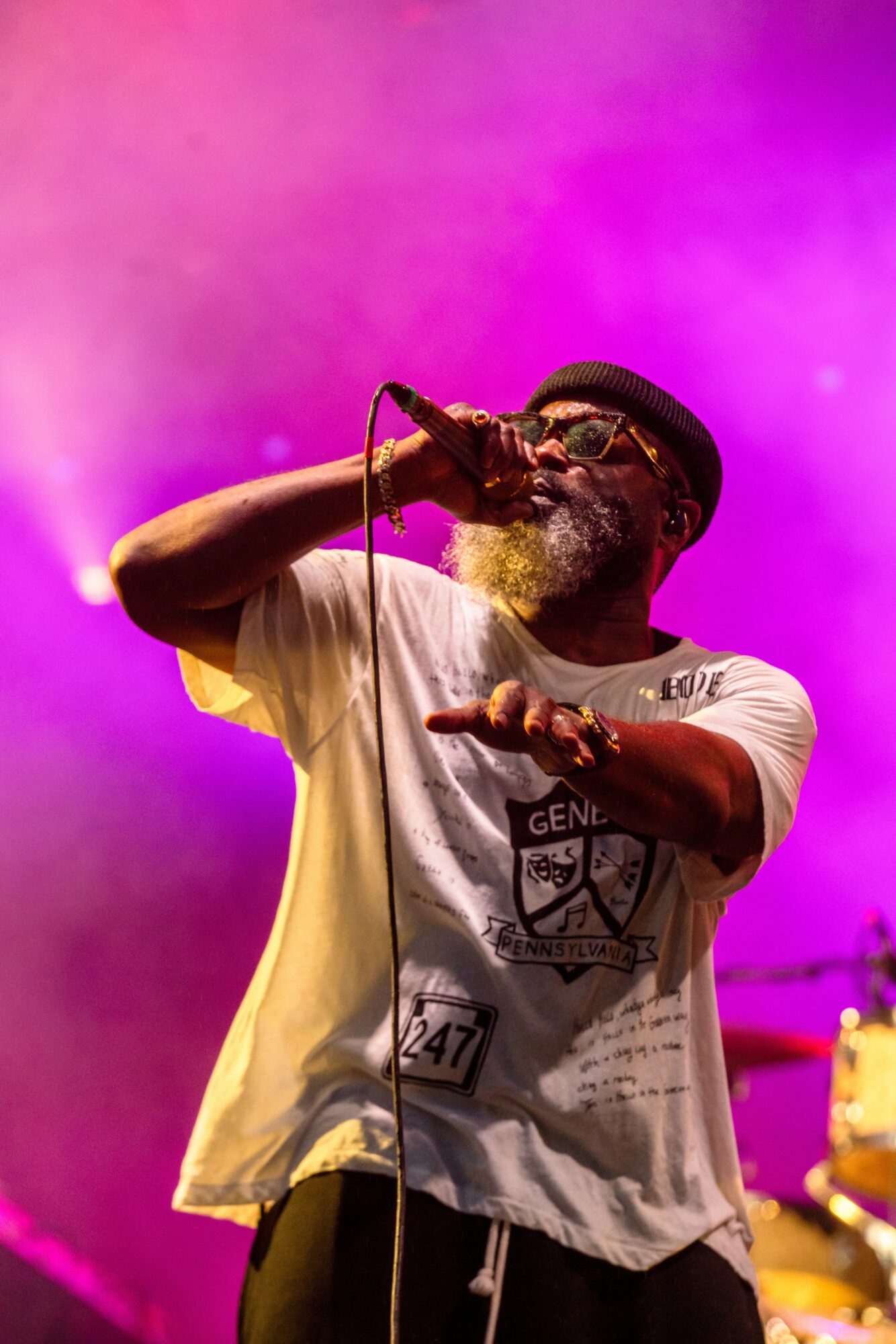 A man singing into a microphone on stage with purple and pink lighting, wearing glasses, a hat, and a white t-shirt.