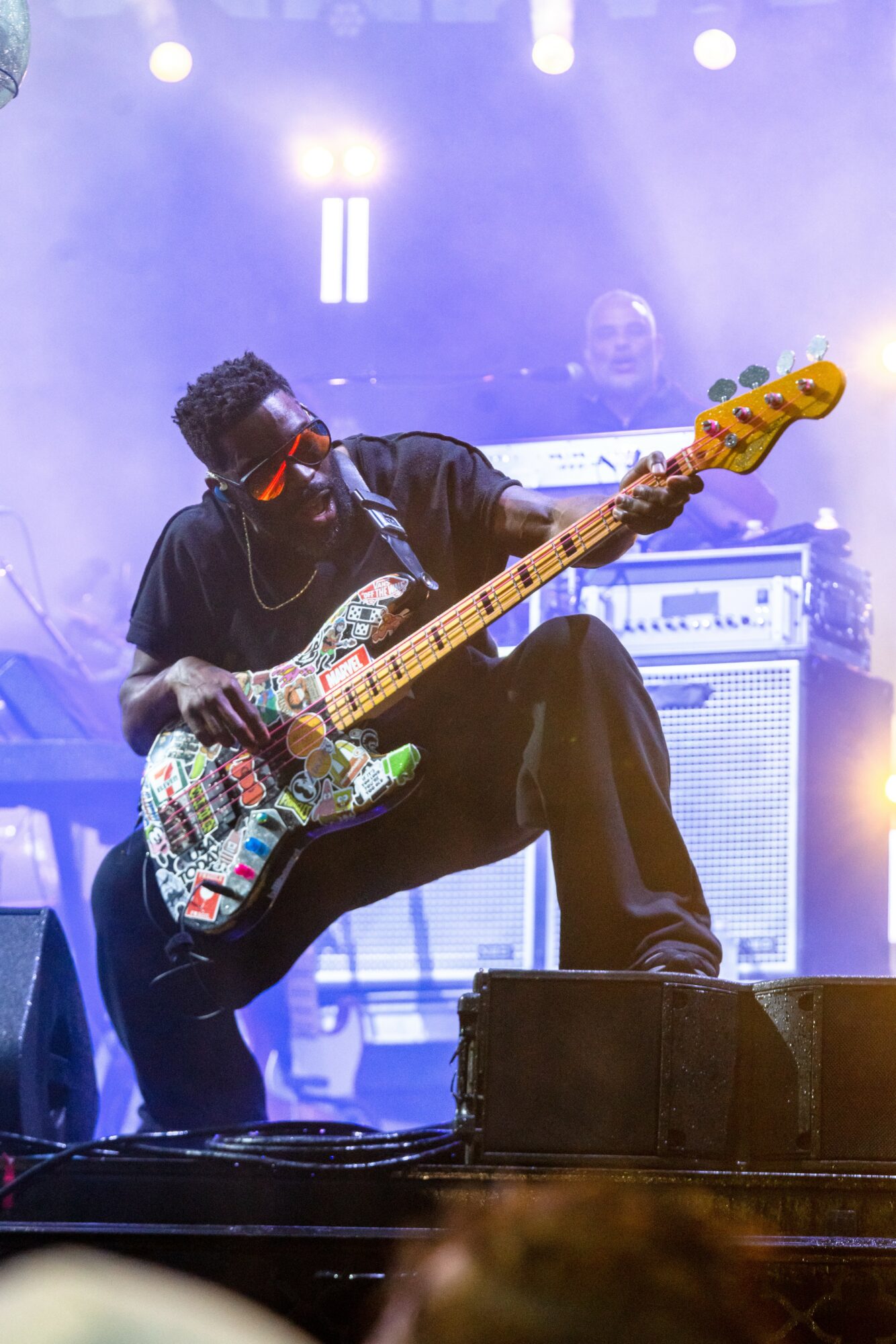 Musician playing a colorful electric bass guitar on stage with bright lights and equipment in background.