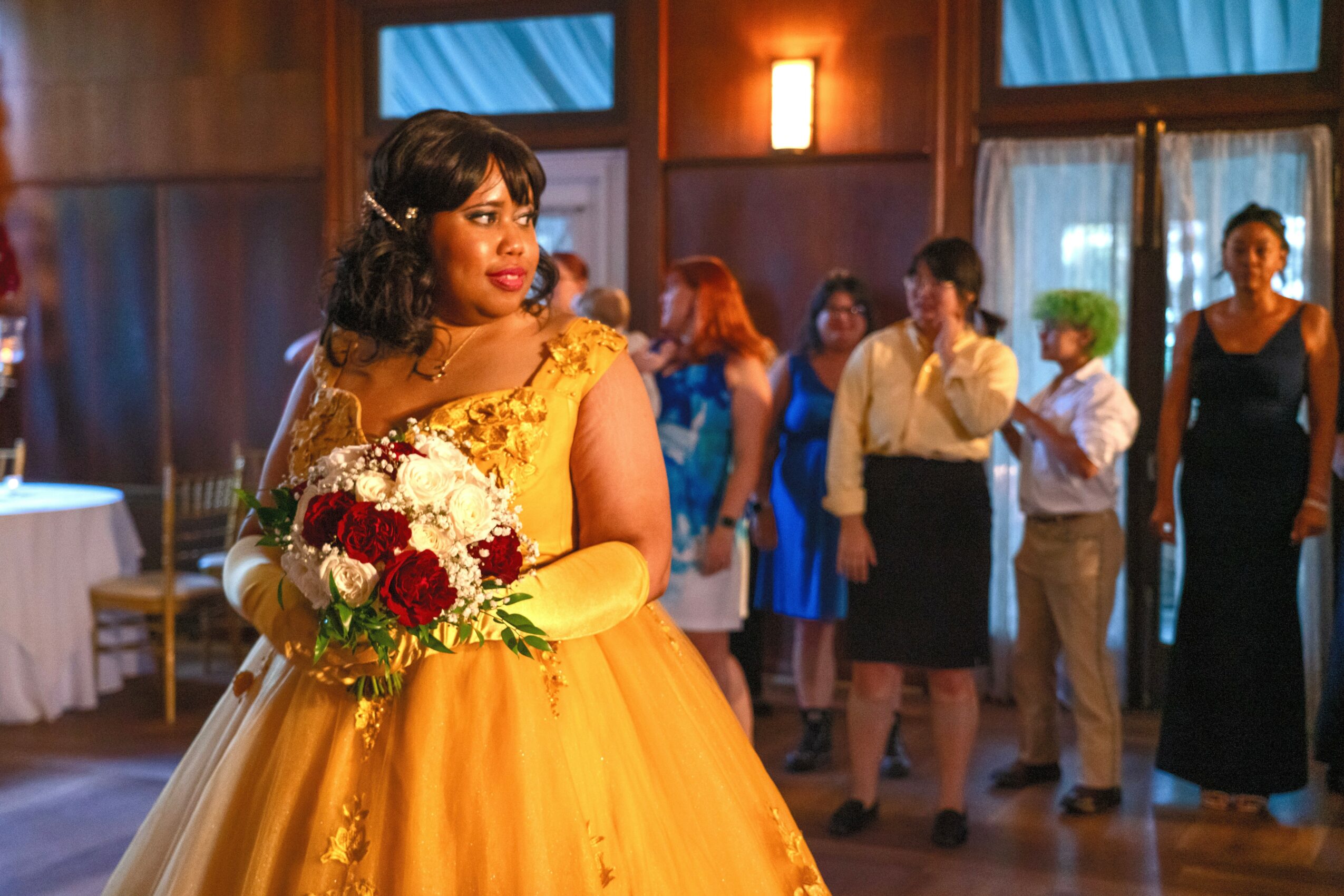 Woman in yellow dress holding a bouquet at a social event with people in the background, indoors with wooden walls and large windows.