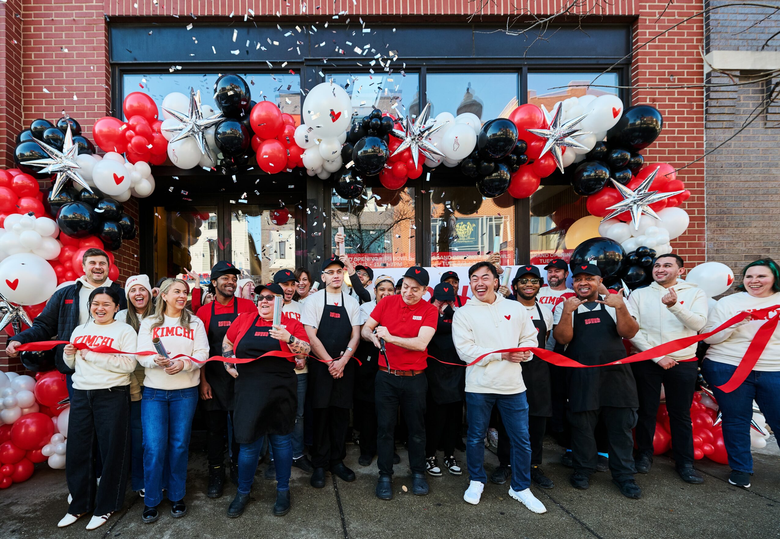Group of people celebrating in front of a decorated storefront with balloons, some holding a ribbon for a ribbon-cutting event.