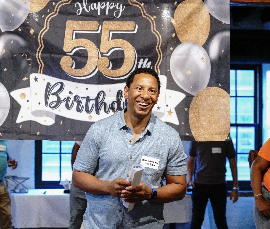Man smiling at birthday celebration with a large decorated banner in the background, holding a paper, in a room with windows.