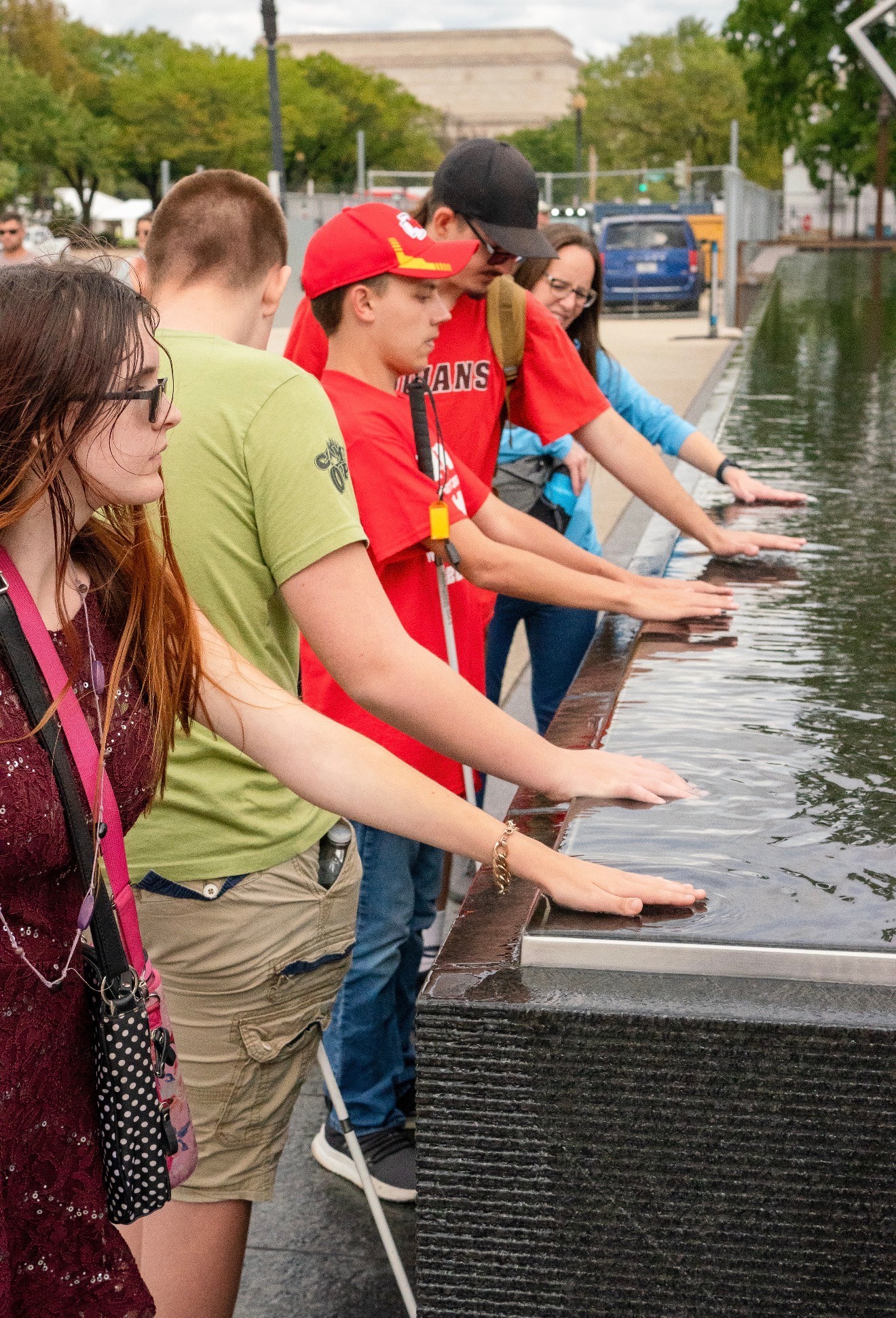 Group of children and teenagers with hands in water at a fountain, outdoors on a sunny day.