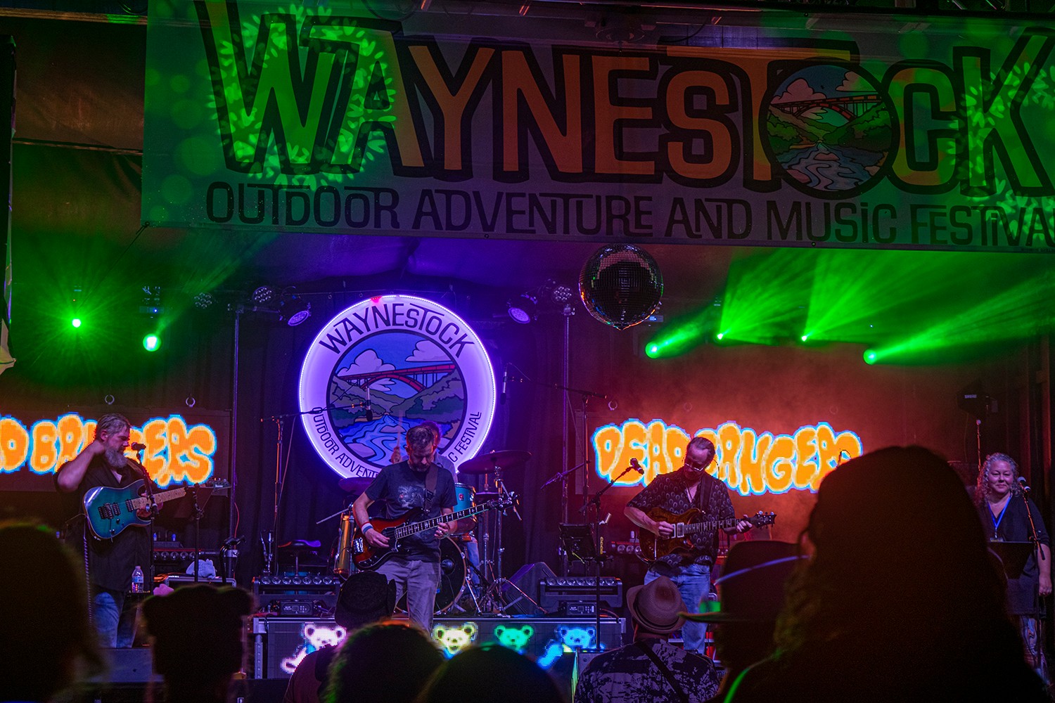 Band performs on stage with colorful background and green stage lights, audience silhouettes in foreground, outdoor music festival setting.