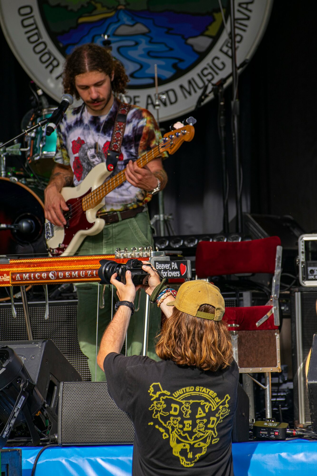 Person playing electric guitar on stage with a large circular emblem in the background, audience member taking photo, stage equipment visible