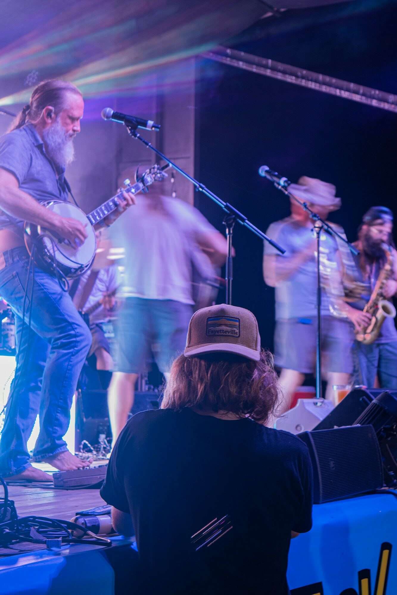 Musicians perform on stage with colorful lighting, including a guitarist, saxophonist, and blurred band members, audience in foreground.