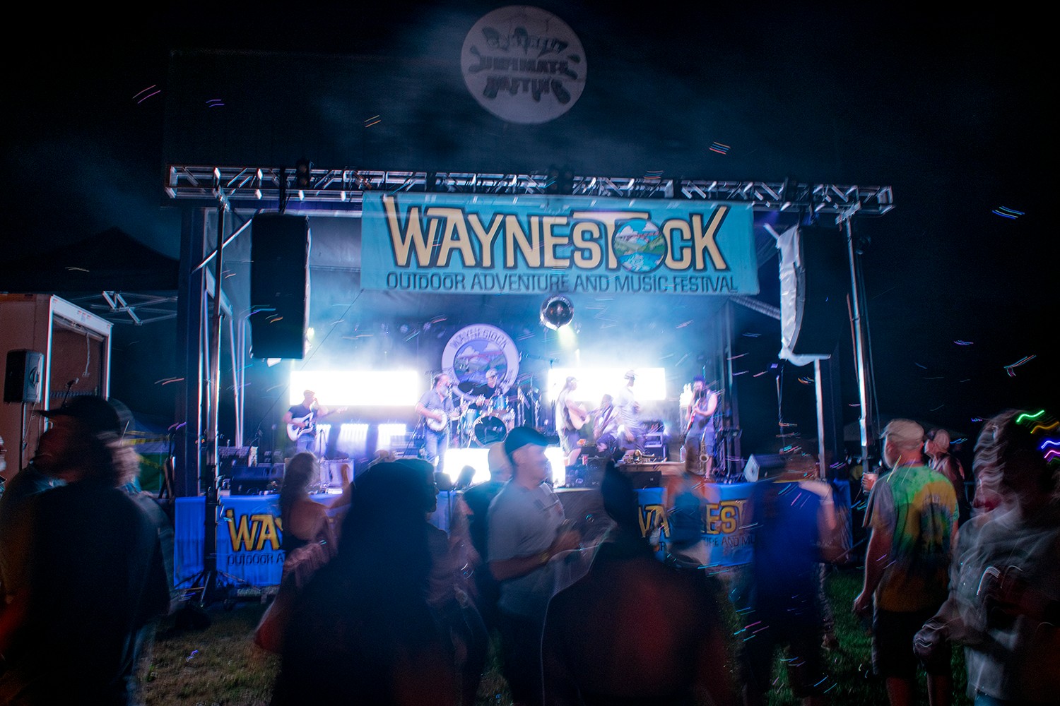 Crowd watching a concert on stage with a large banner reading 'WAYNESC' and bright lights, indoors or at night.