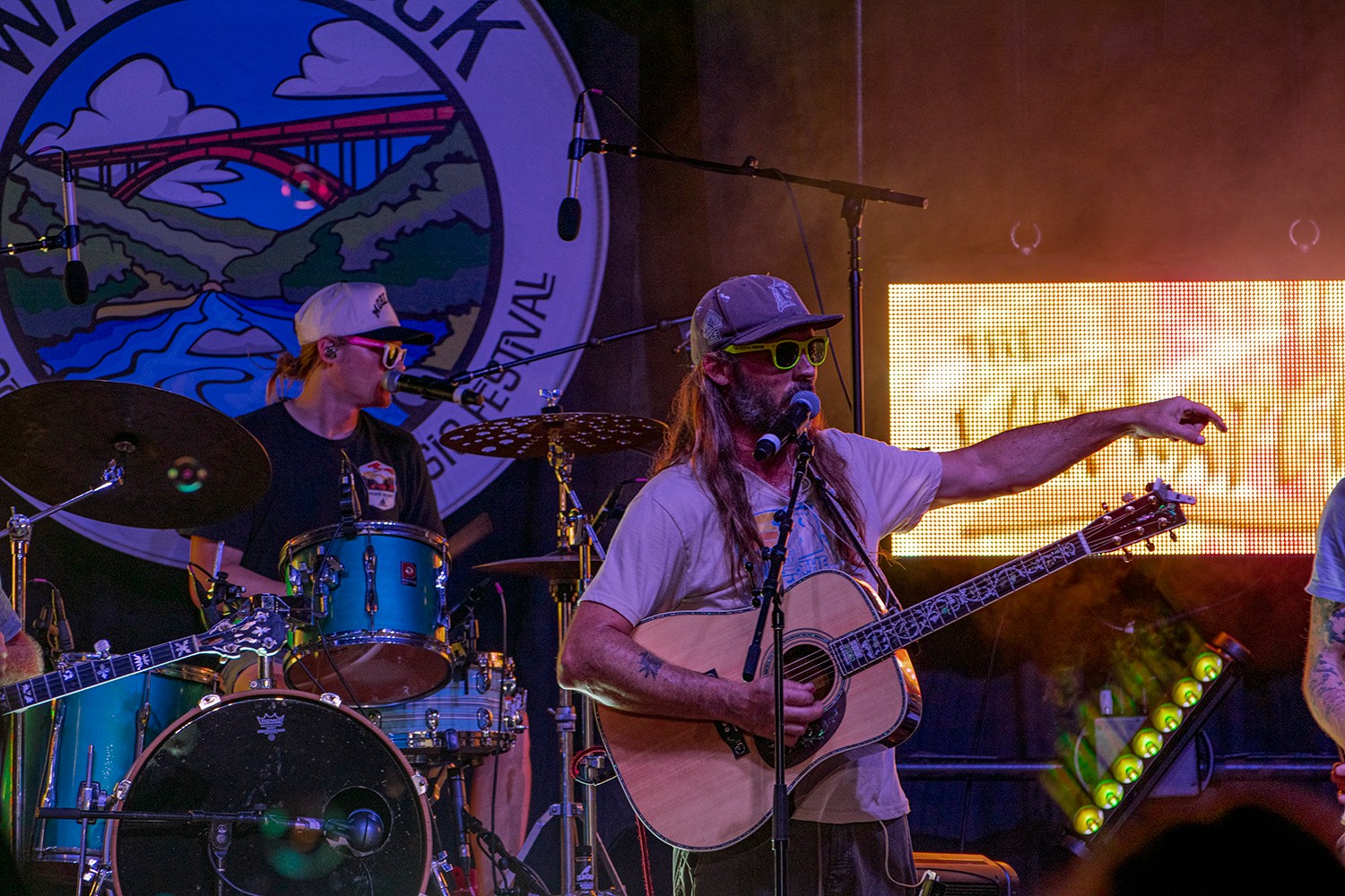 Two musicians perform on stage, one playing drums and the other singing and playing guitar, with a festival logo in the background.
