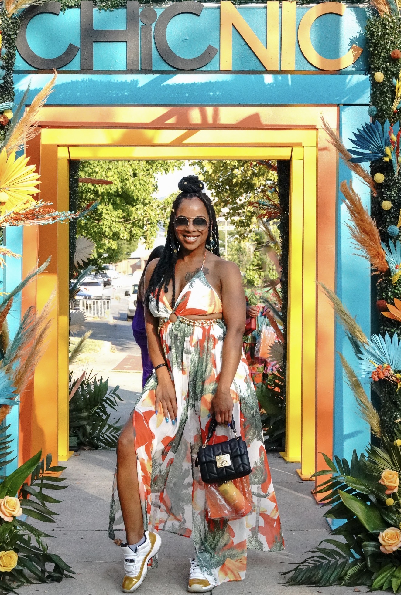 Woman in a colorful dress standing in a decorated archway outdoors, holding a handbag and smiling.