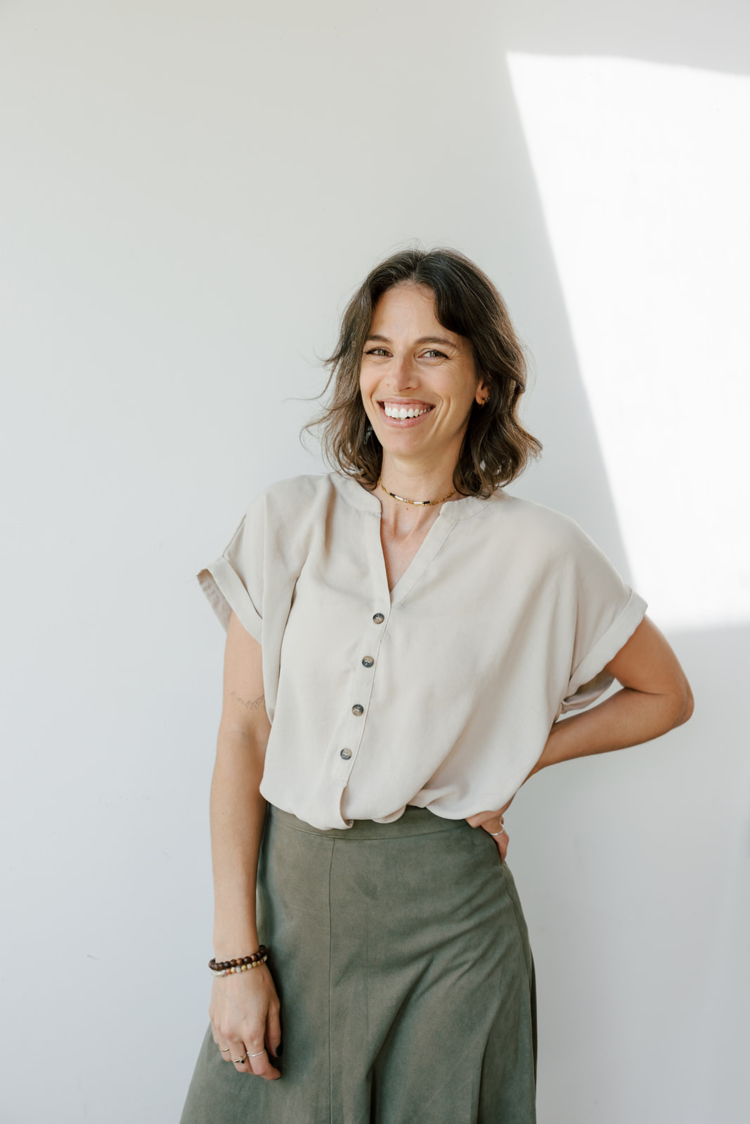 Woman smiling, standing with hand on hip, wearing a light-colored blouse and gray skirt, in bright room.