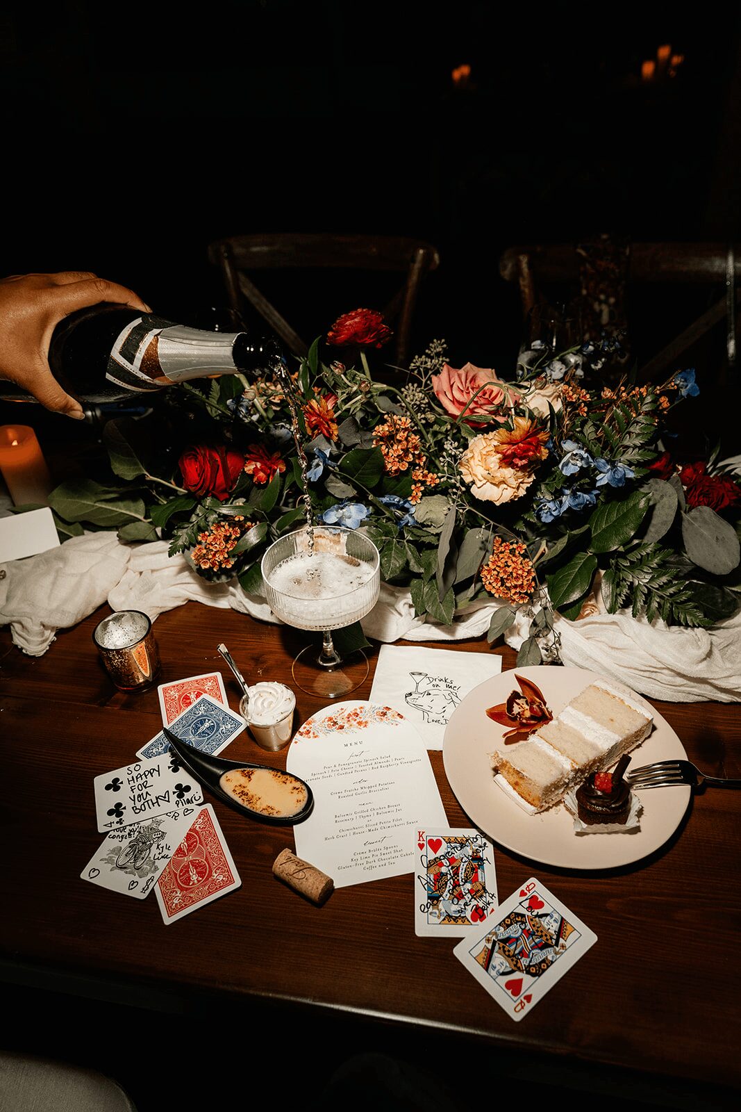 Table with flowers, drinks, playing cards, and desserts in a dimly lit setting.