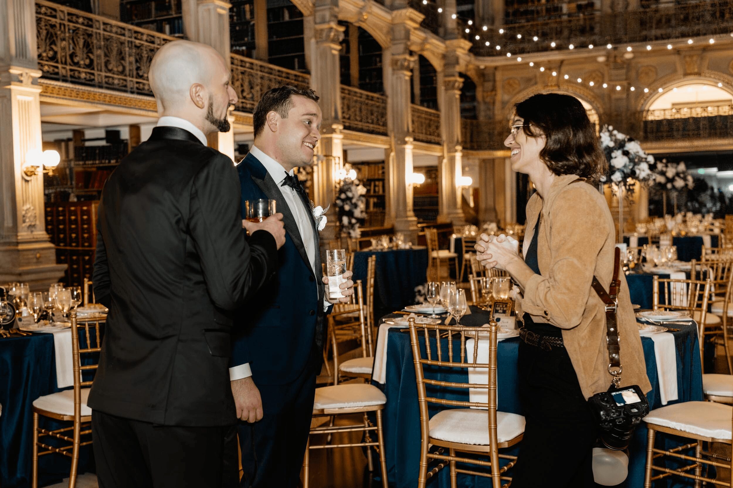 Three people engaged in conversation at a formal event, with decorated tables and elegant interior in the background.