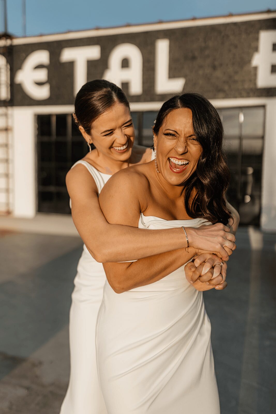 Two women in white dresses laughing and hugging outdoors in front of a building with large windows and partial text.