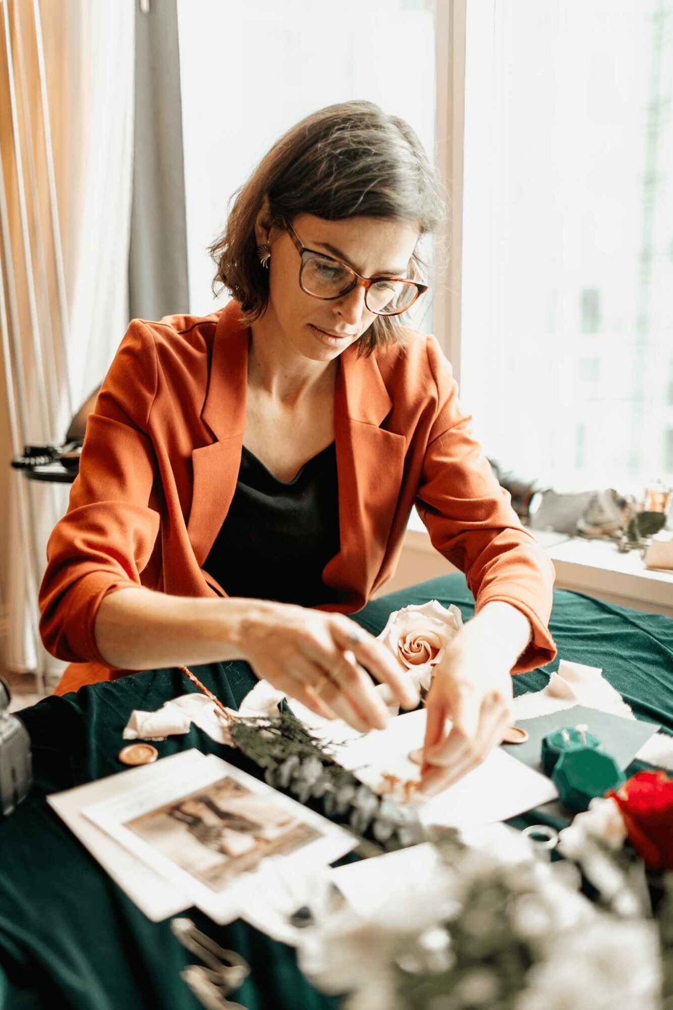 Woman with glasses arranging flowers at a table, sunlight coming through window, workspace with papers and flowers.