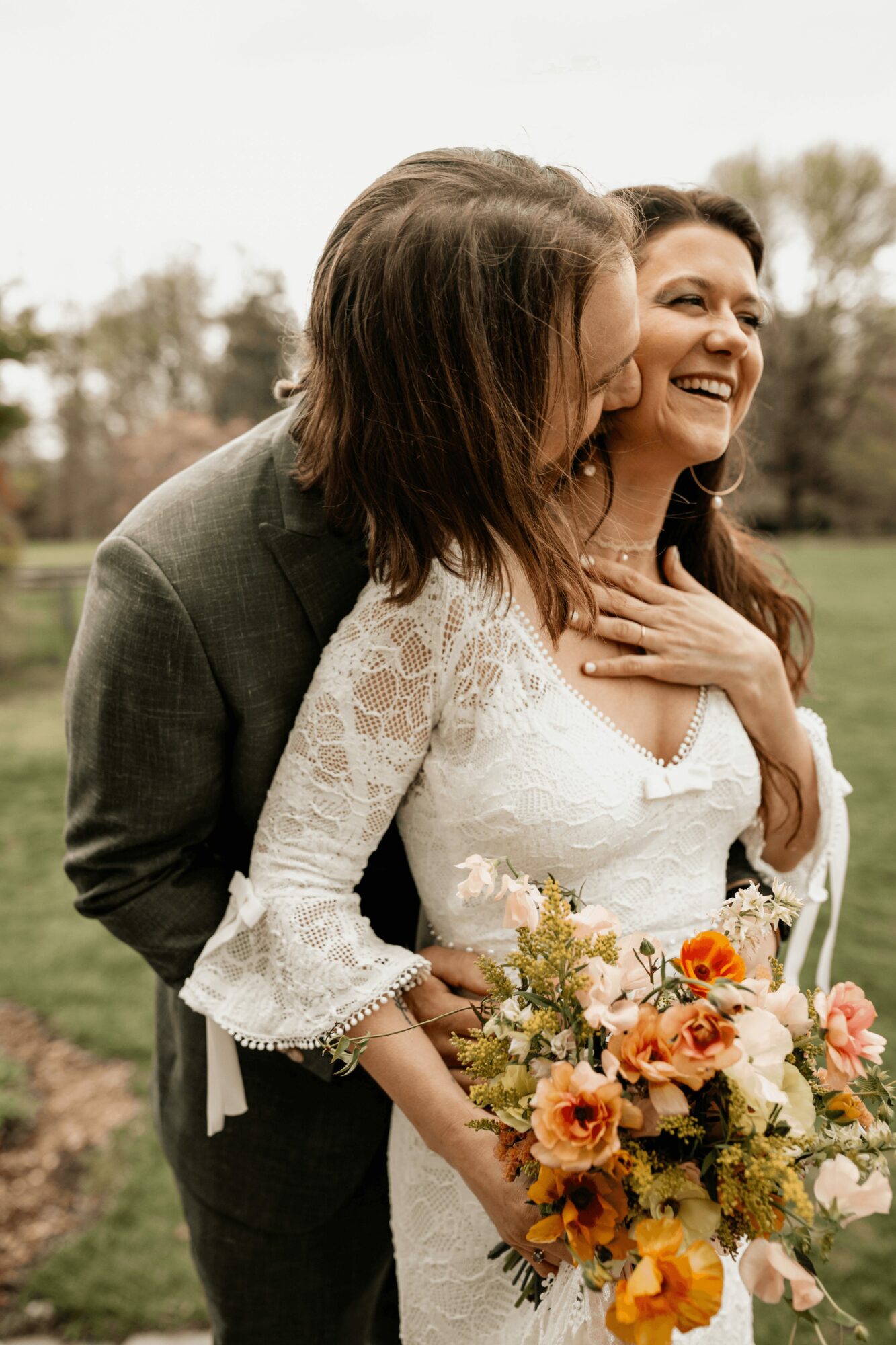 Two women, one in a wedding dress holding a bouquet, smiling outdoors with trees in the background.