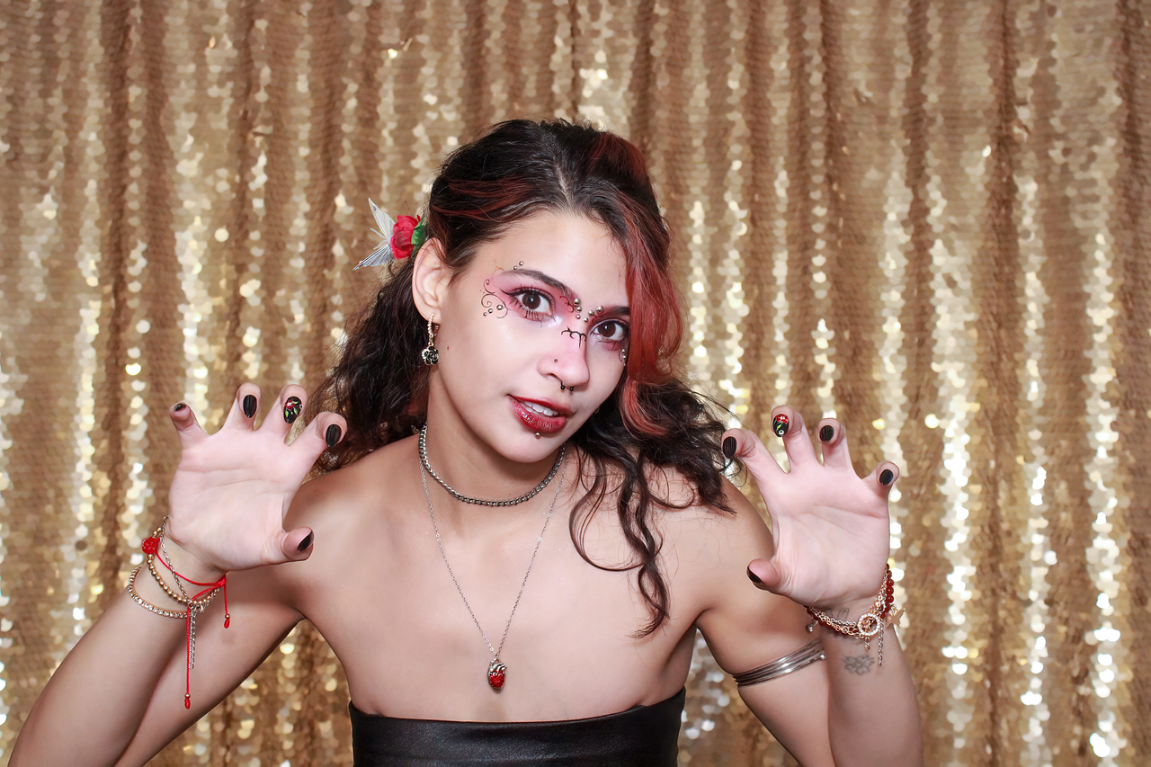 Young woman with makeup and jewelry poses with hands raised, against a gold sequin backdrop.