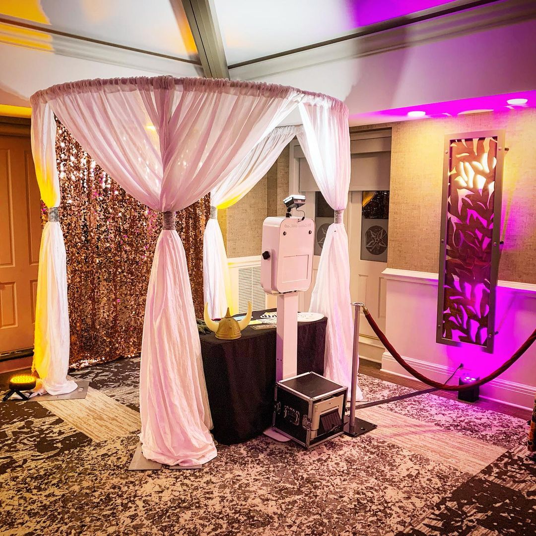 Photo of a photo booth with pink curtains, a sequin backdrop, and a camera on a stand, in a decorated indoor setting.