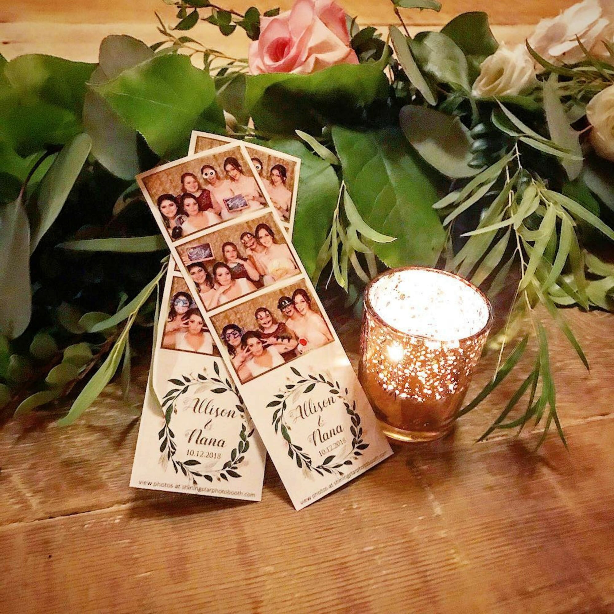 Photo booth strip with four pictures, two place cards with floral designs, a lit candle, and flowers and greenery on a wooden surface.