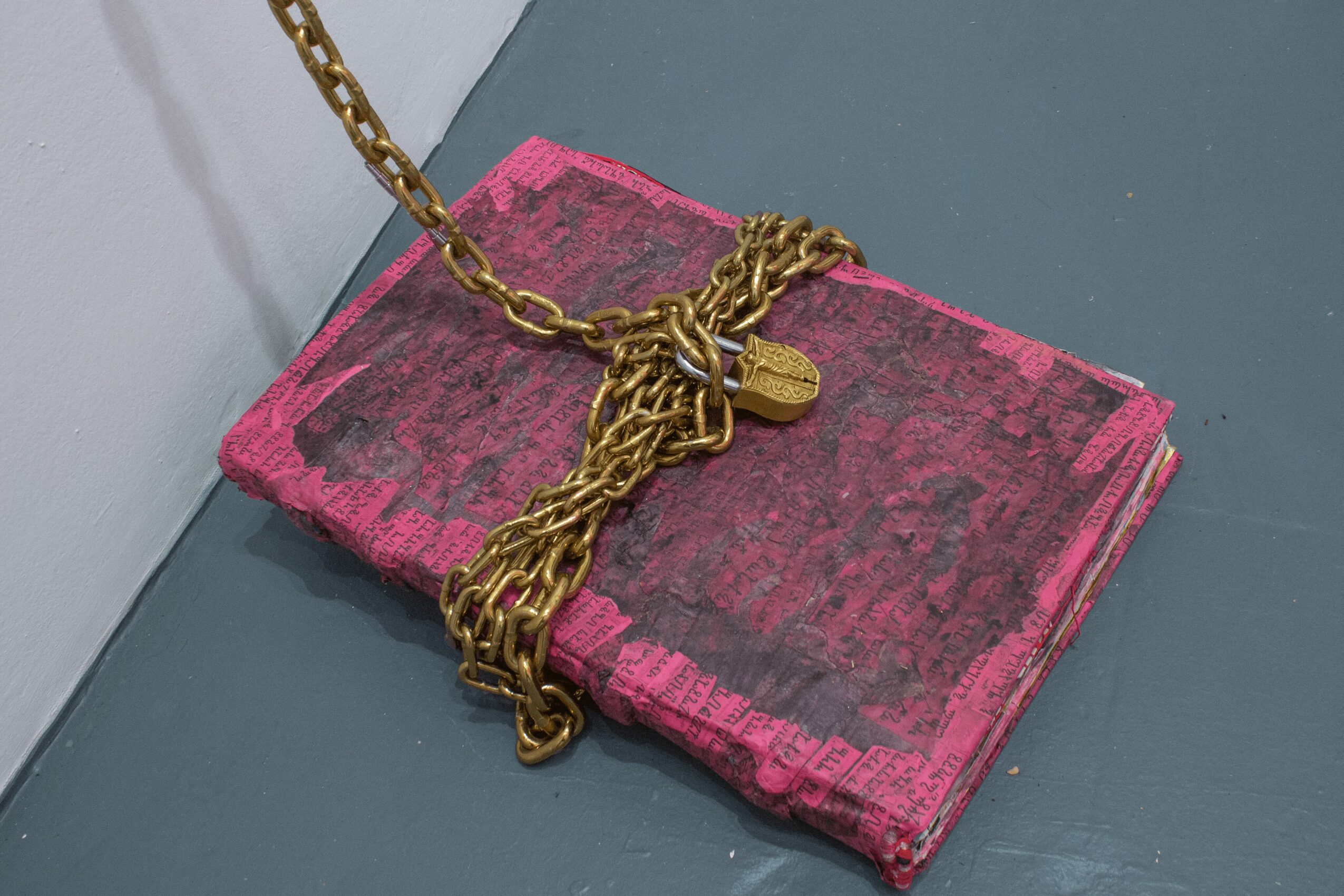 Pink textured clutch with a gold chain and small gold tag, placed on a gray surface.
