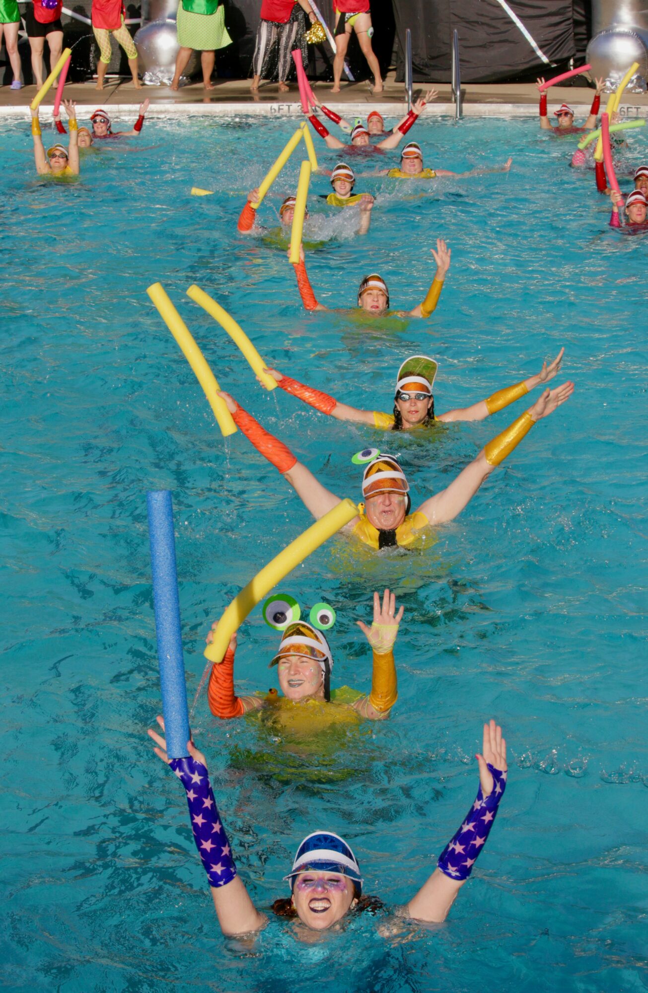 Swimmers in a pool participate in a water activity with colorful foam noodles, some waving and smiling, with spectators on the poolside.