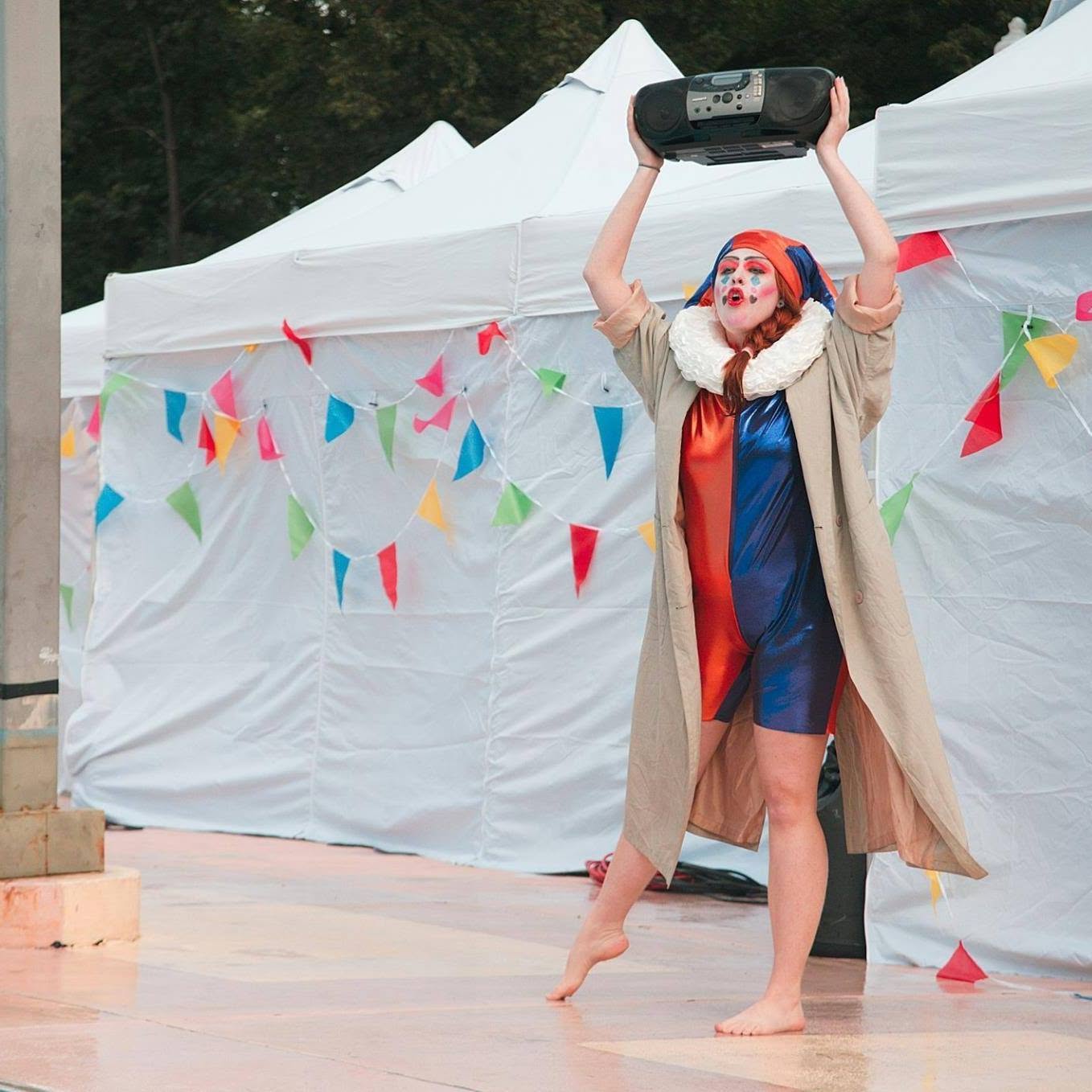 Clown with face paint and costume holding a boombox above head, standing barefoot outdoors near white tents and colorful decorations.