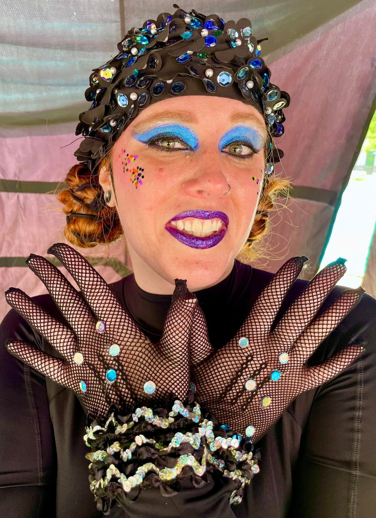 Person with colorful makeup, wearing a beaded headpiece, fishnet gloves, and a black outfit, smiling with crossed arms.
