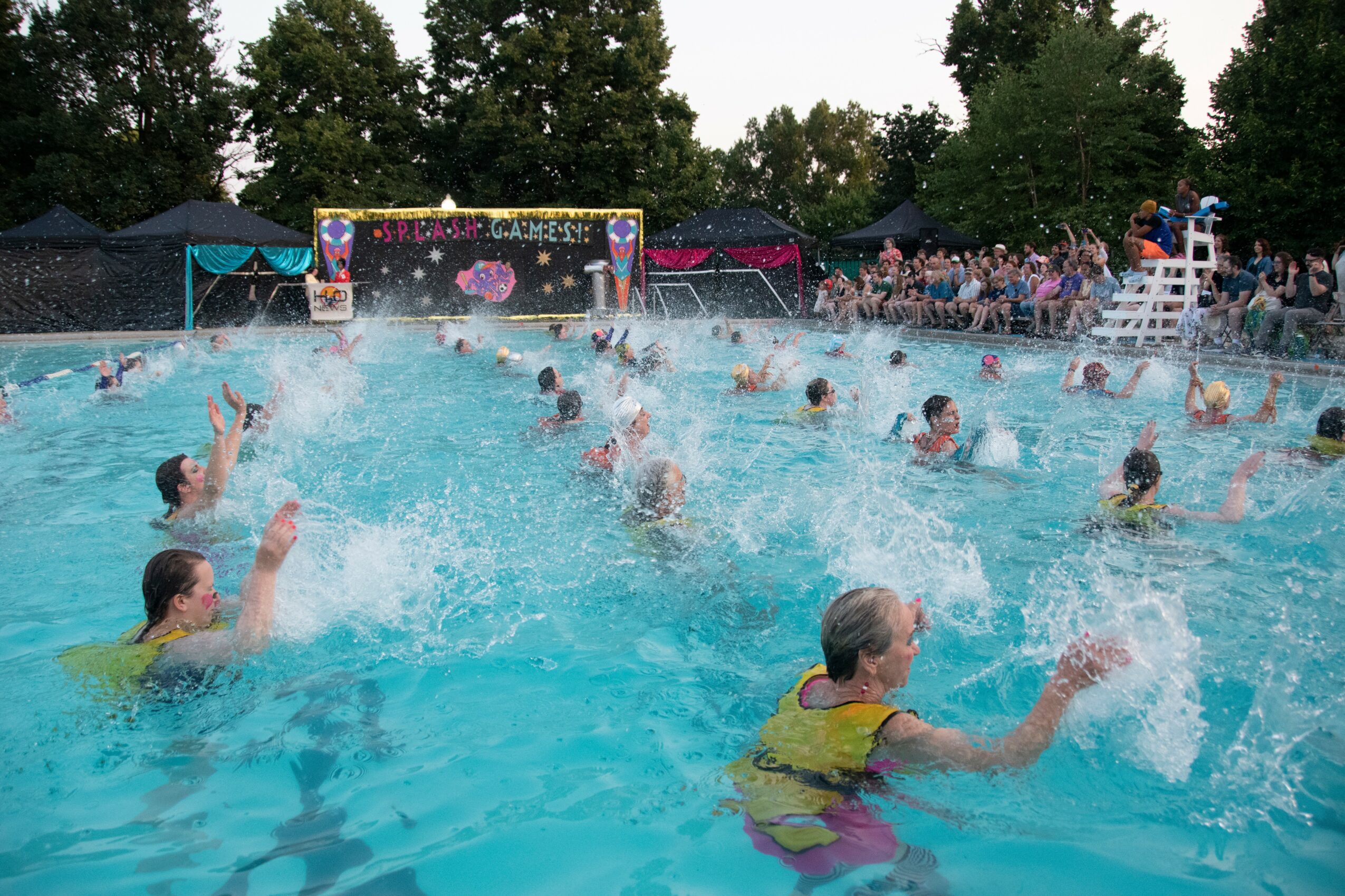 People playing water volleyball in a swimming pool with spectators on the side, trees in the background.