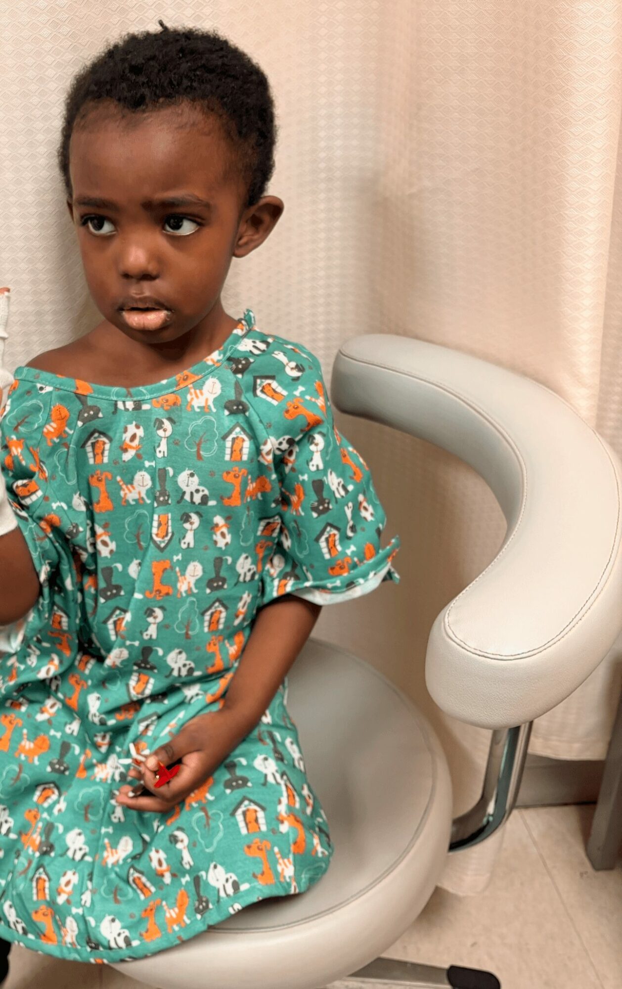 Young girl with curly hair sitting on a medical examination chair, wearing a colorful dress with animal patterns, looking to the side.