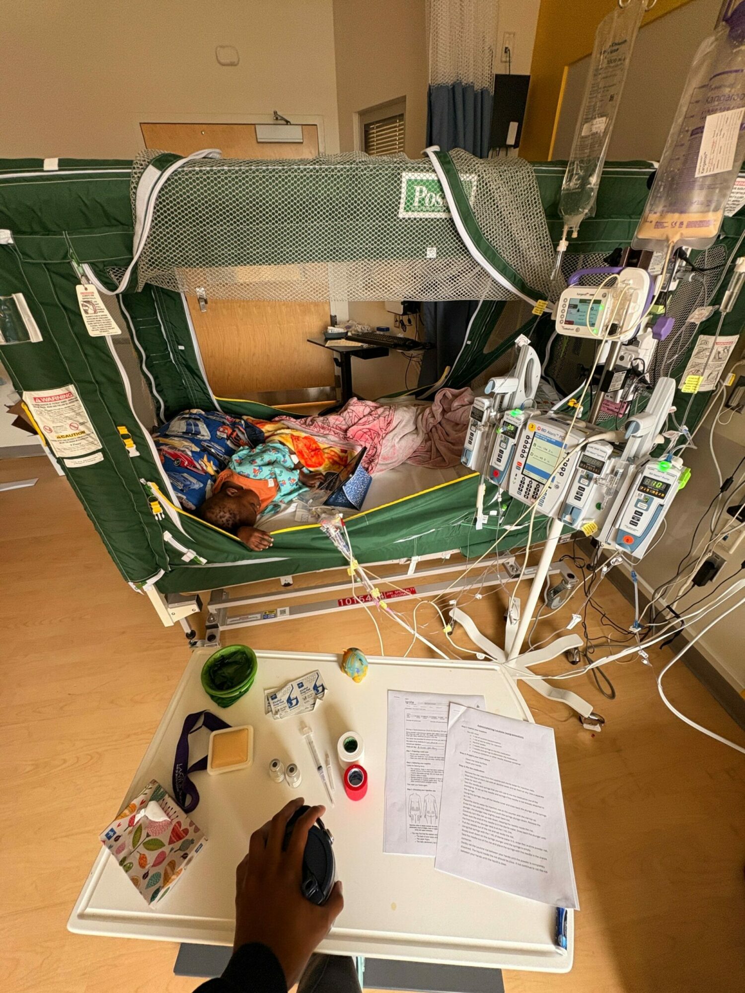 Medical equipment connected to a patient lying on a hospital bed, with a healthcare worker's hand nearby, and documents on a table.
