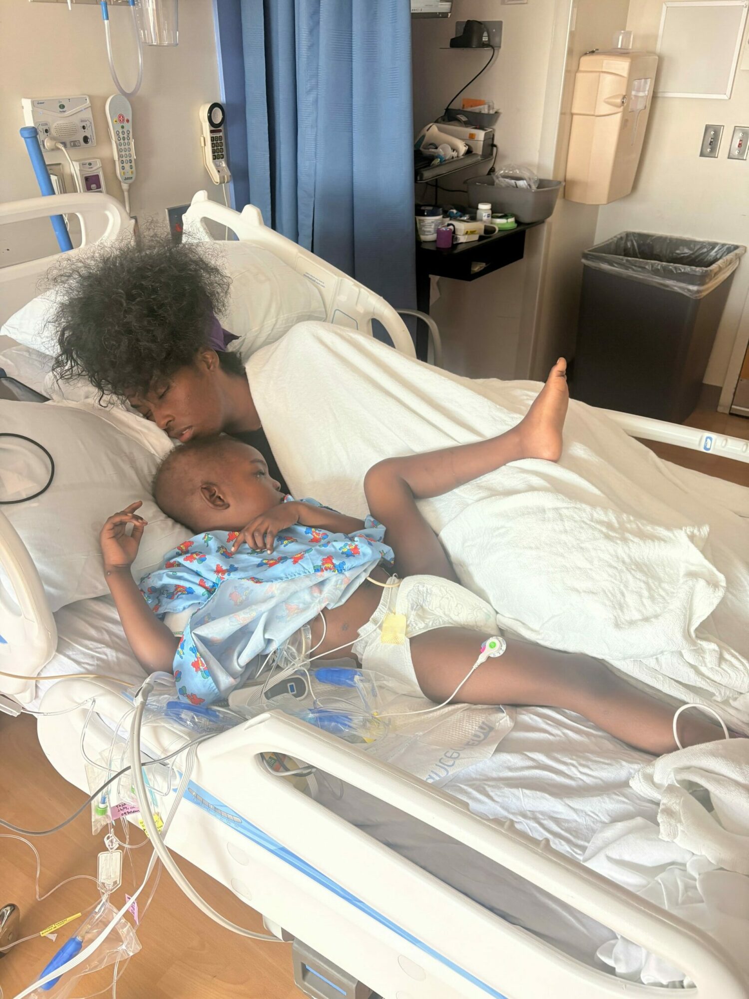 Two infants lying in hospital beds, one with curly hair and the other with a shaved head, connected to medical equipment.