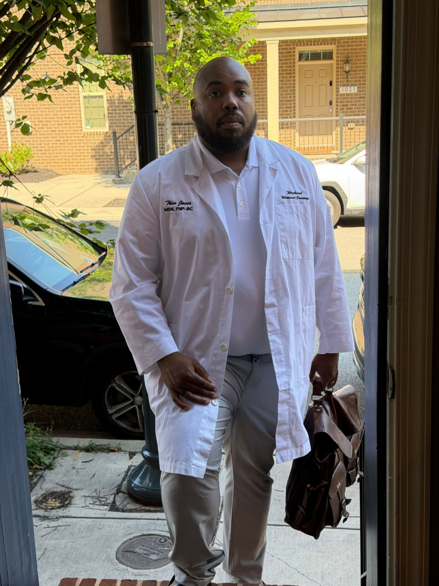 Person in white coat standing outside near parked cars, holding a phone, with buildings and trees in background.