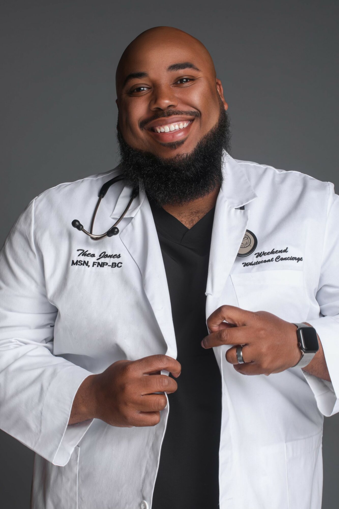 Smiling man in white medical coat with stethoscope, standing against gray background.