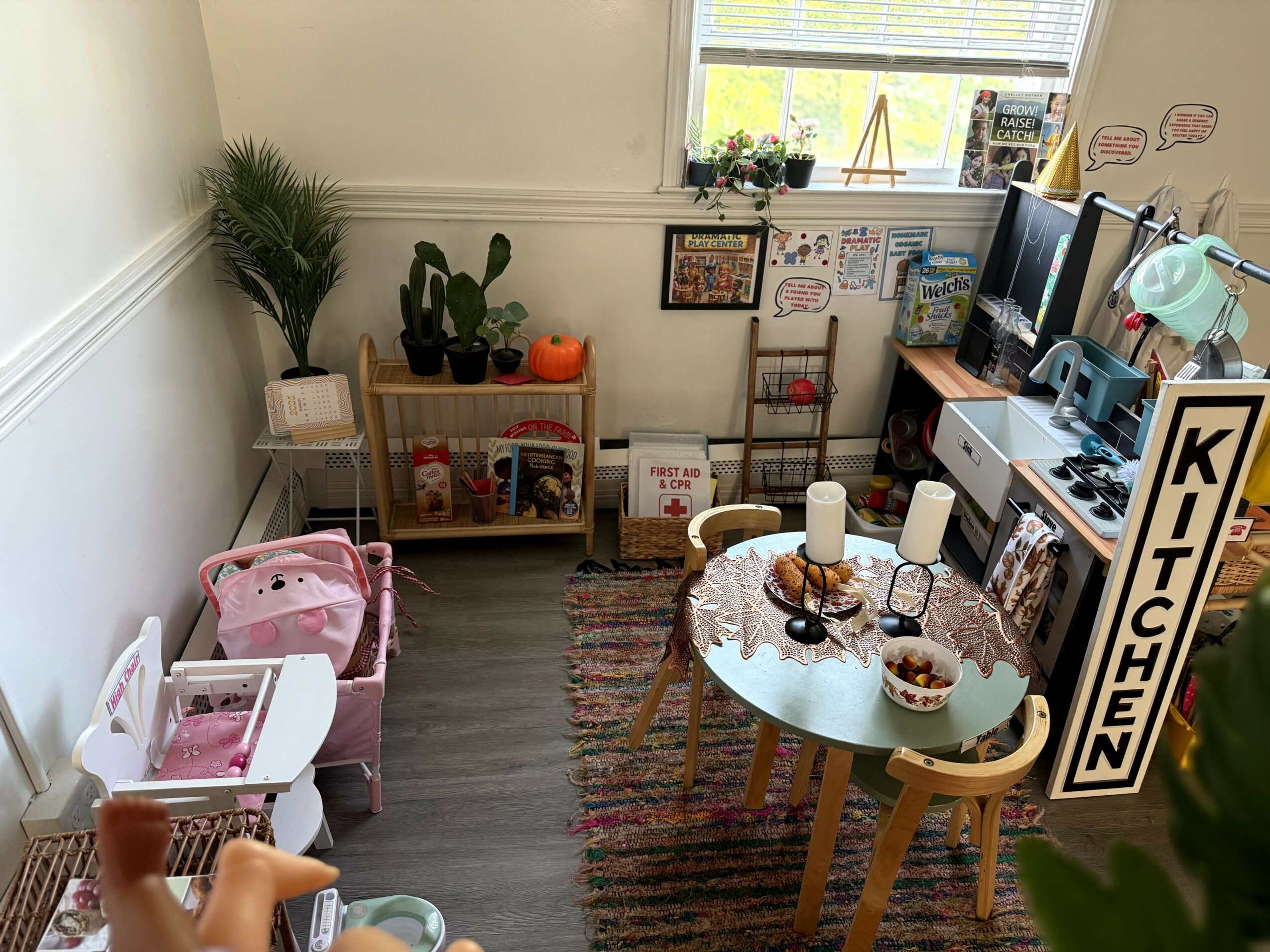 Room with a round table, chairs, plants, and a window, decorated with children's drawings and toys, and a kitchen sign.