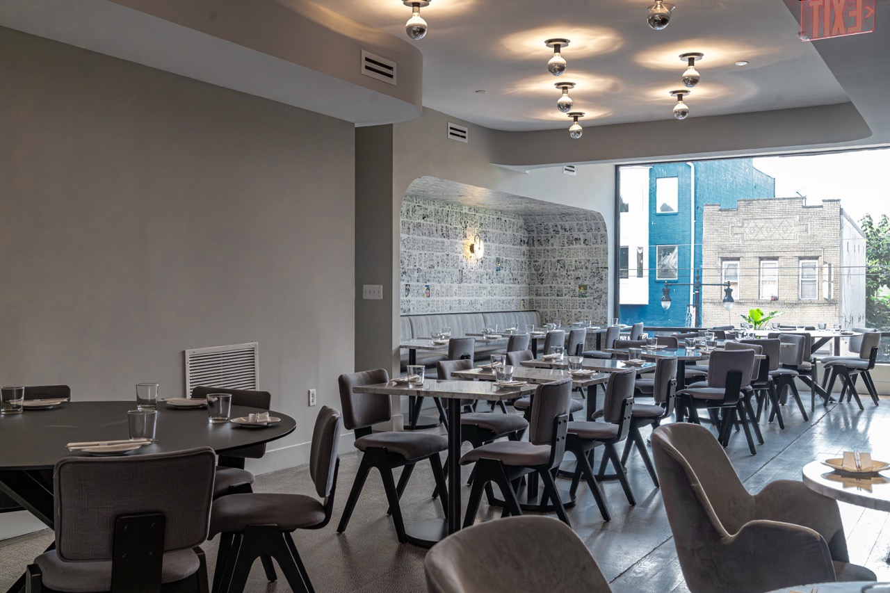 Empty restaurant with tables and chairs, large window, modern decor, and ceiling lights, daytime setting.