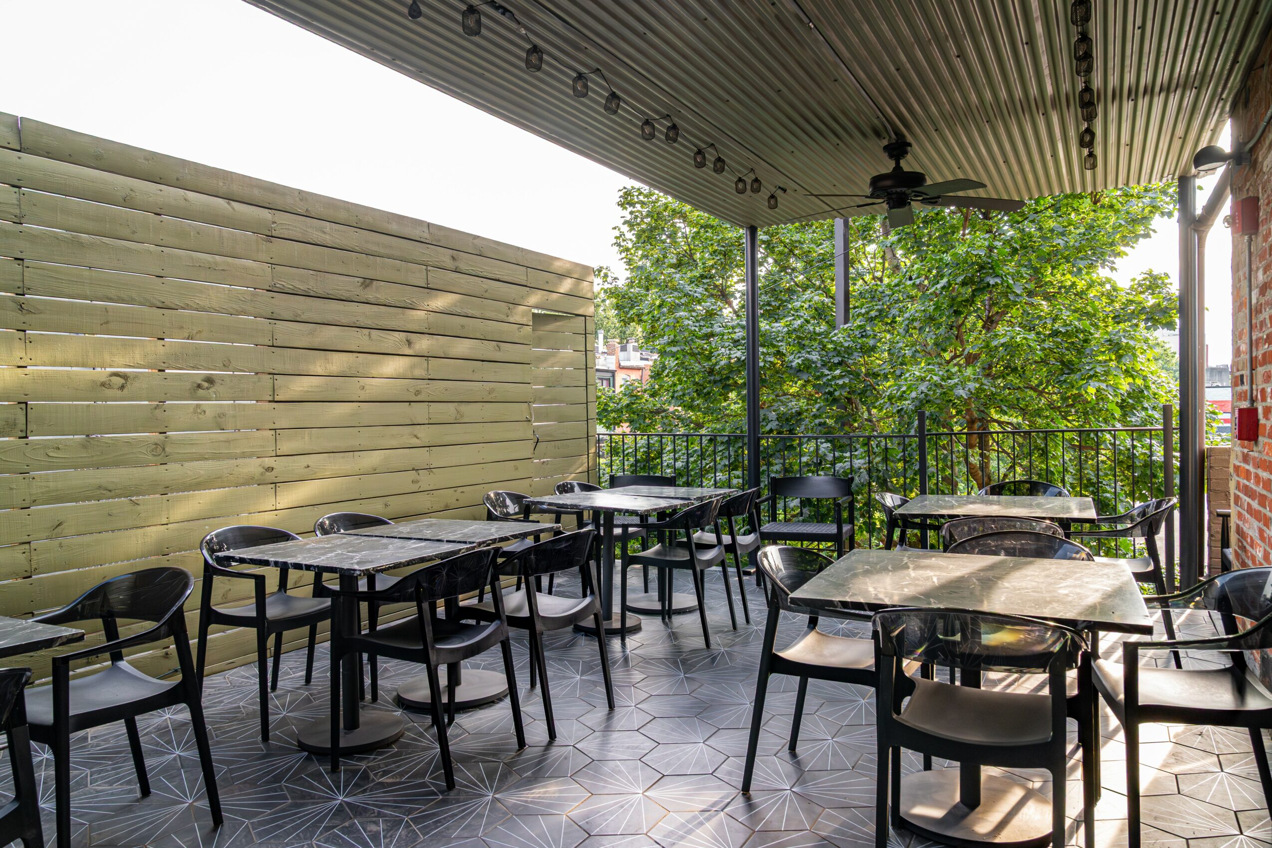 Outdoor dining area with tables and chairs, surrounded by a wooden fence and greenery, under a covered roof.