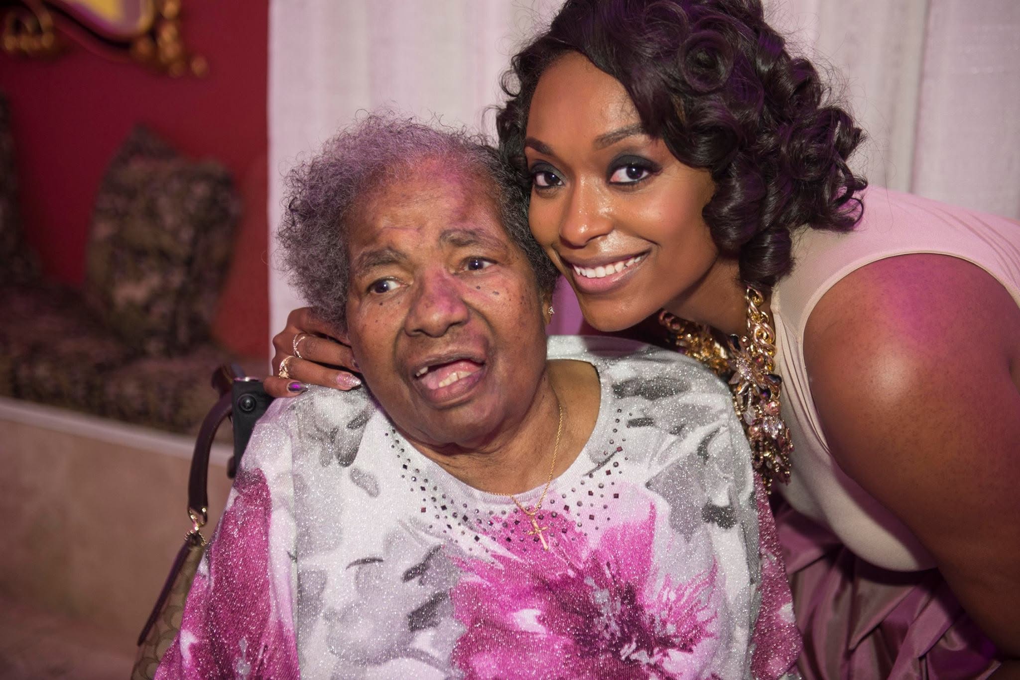 A young woman with curly hair smiling next to an elderly woman with gray hair, both indoors.