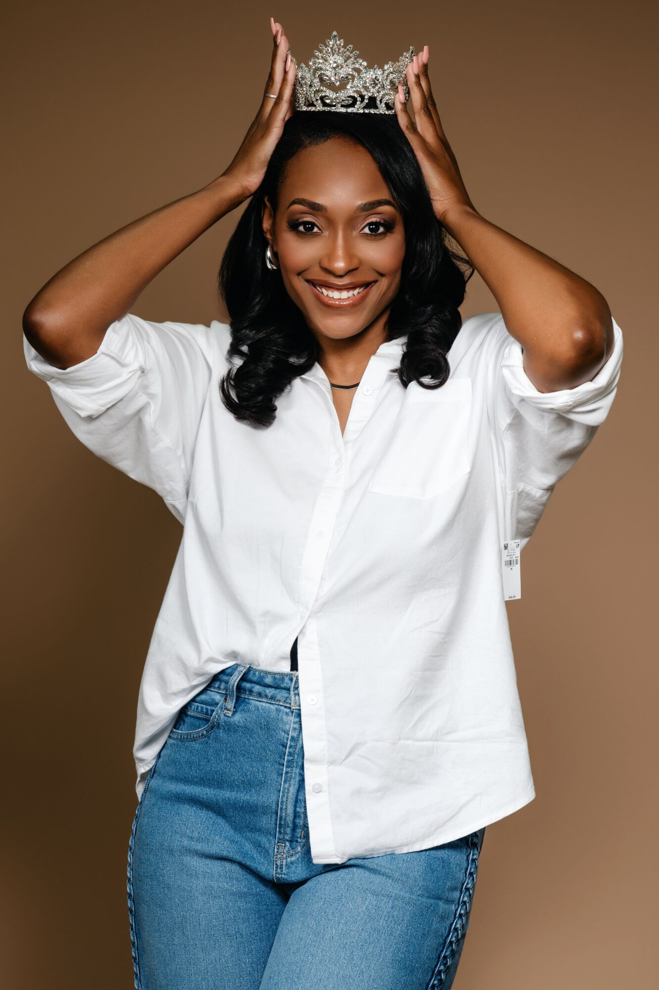 Woman wearing a white shirt and blue jeans, holding a crown on her head, smiling, against a brown background.