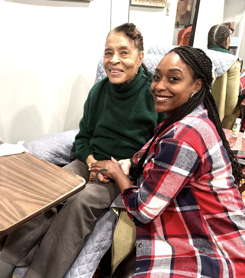 Two women smiling, one sitting in a chair and the other standing beside her, in a room with mirrors and furniture.