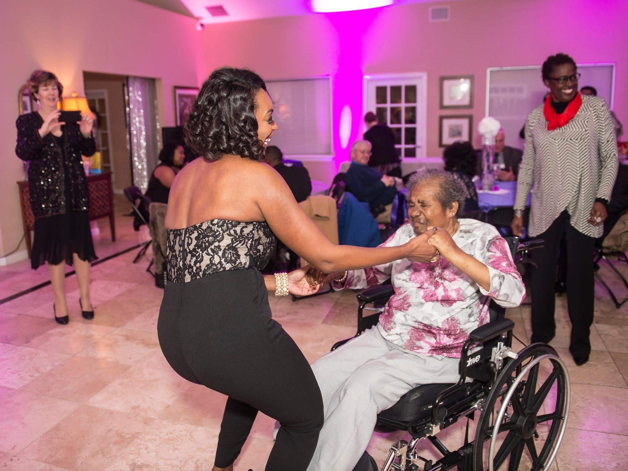 Woman in black dress interacts with woman in wheelchair at a social event, others in background, pink lighting, indoor setting.