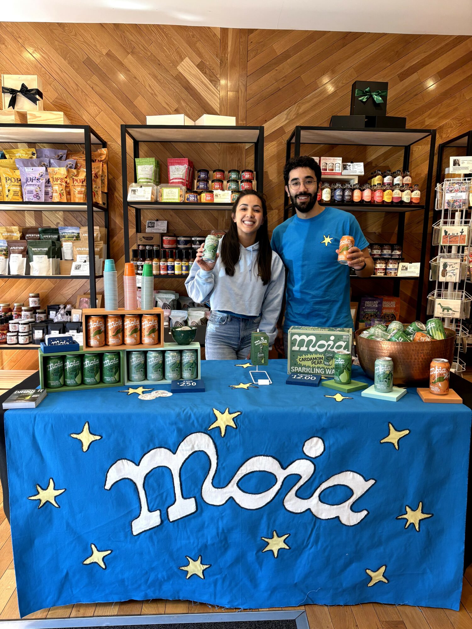 Two people standing behind a table with a blue cloth and the word 'Moia' in a store, shelves with products behind them.