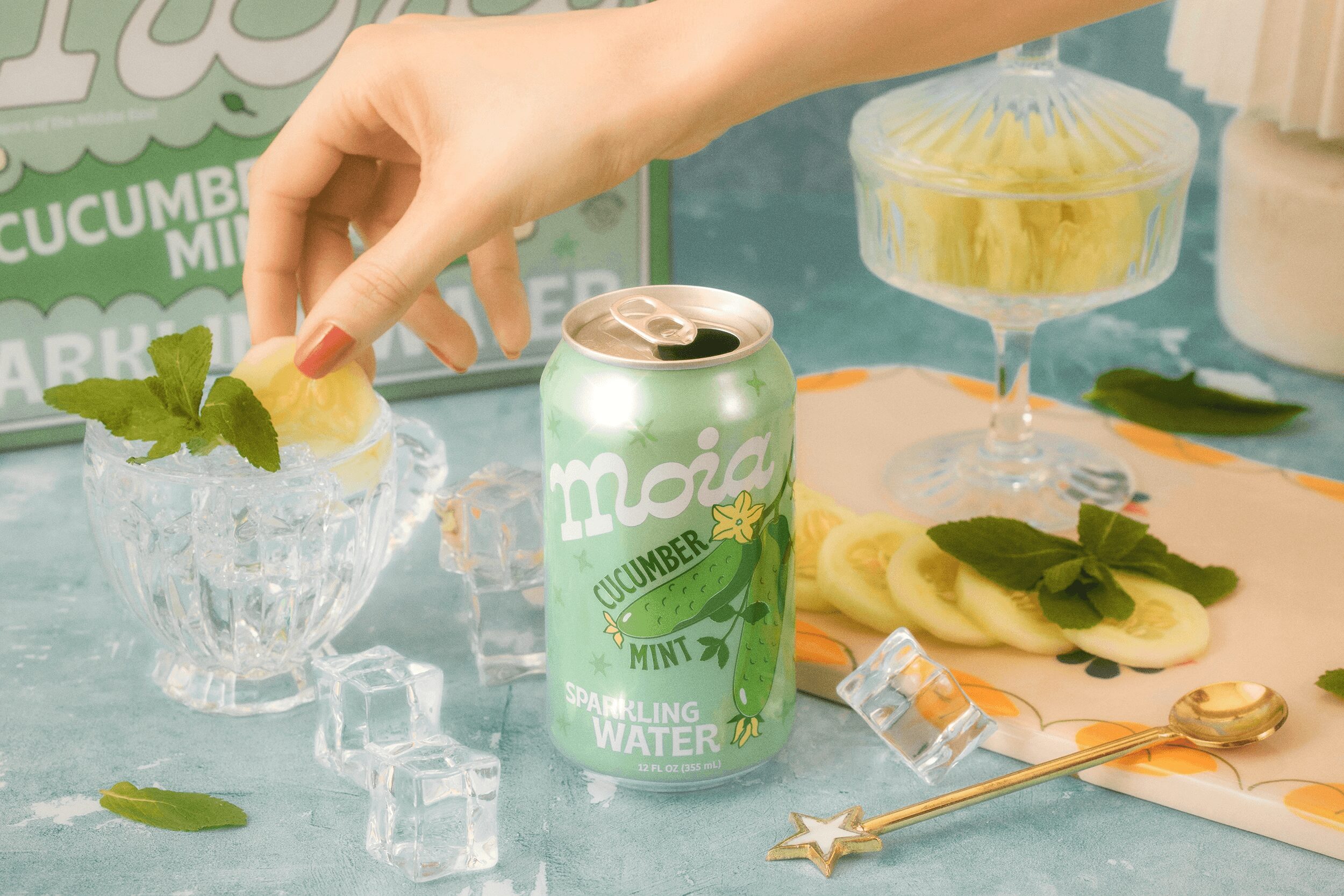 Hand placing lemon slice and mint into a glass with ice, with a can of cucumber mint sparkling water nearby, on a table.