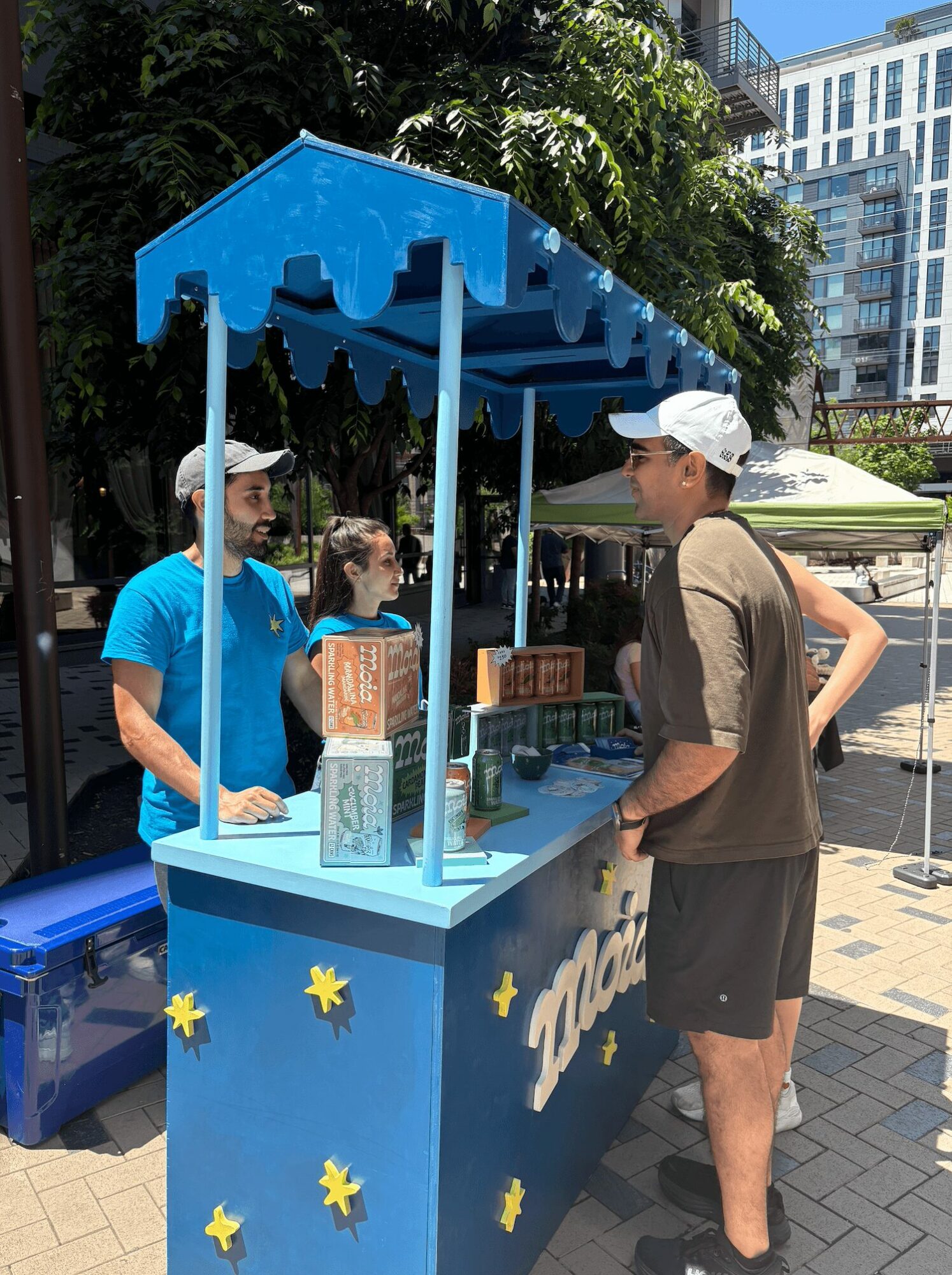 A man and a woman stand at a blue ice cream stand with a canopy, surrounded by trees and buildings.