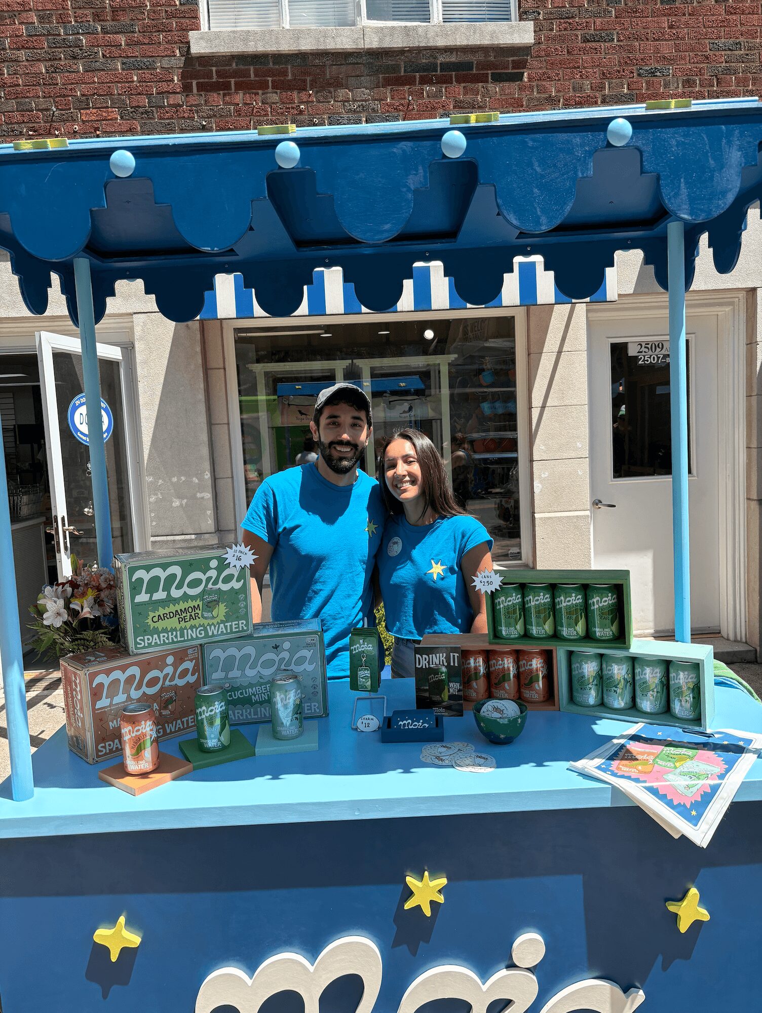 Two people stand behind a blue booth with drinks and a sign, smiling outdoors in front of a building.