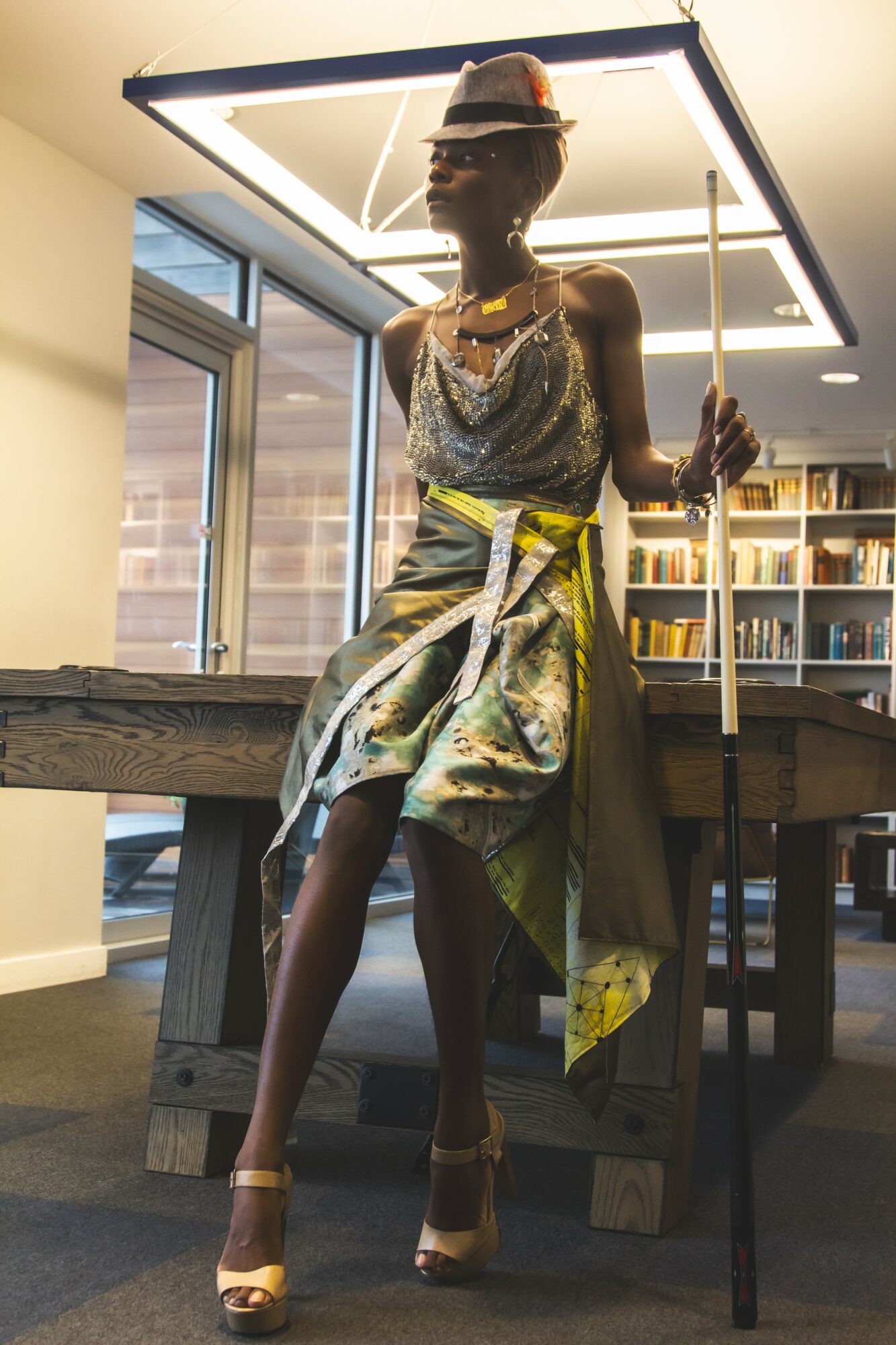 Fashionable mannequin in a metallic dress, hat, and jewelry, holding a staff, standing in a room with bookshelves and windows.
