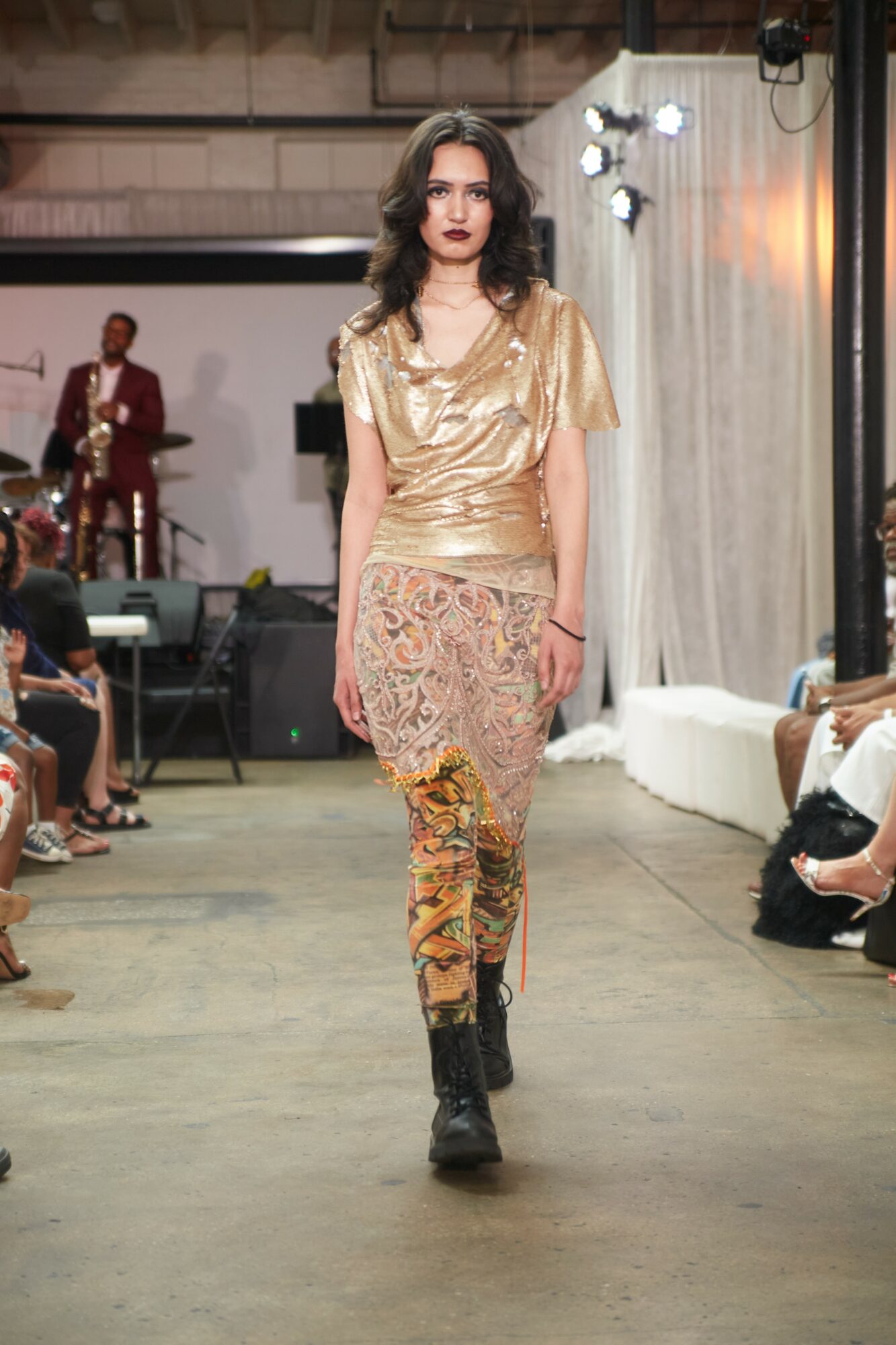 Female model walking down runway in gold top and patterned pants, audience seated on sides, stage with musicians in background.
