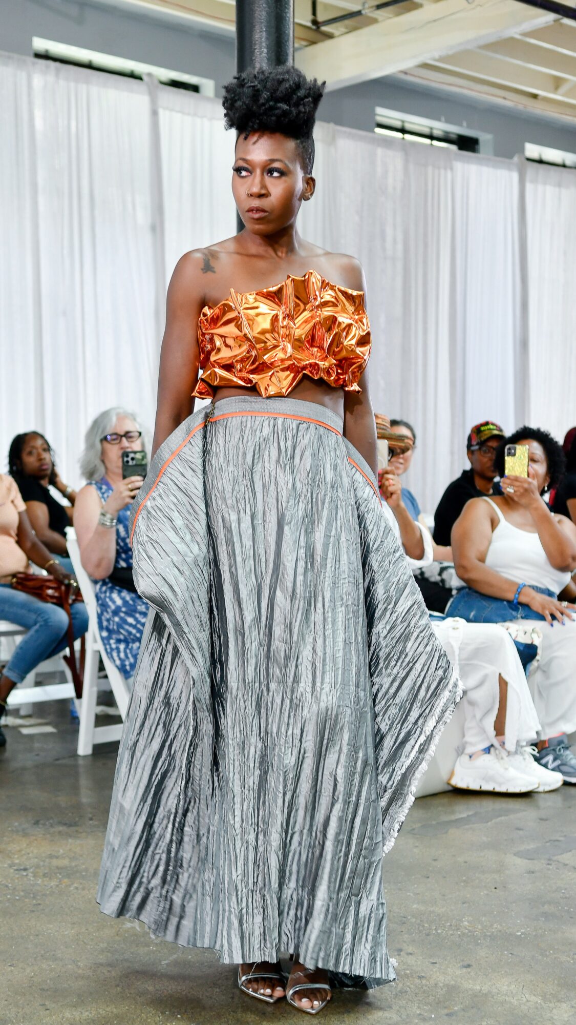 Woman walking on runway wearing an orange ruffled top and a long, striped gray skirt, audience seated in background.