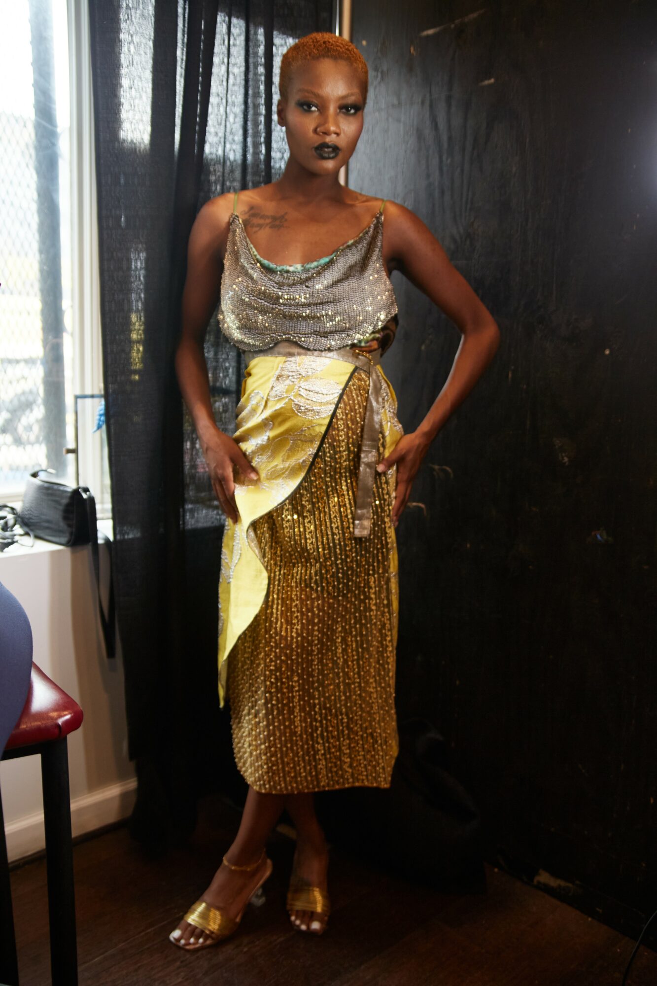 Woman with short hair in a silver top and patterned skirt, standing indoors near a window and black wall.